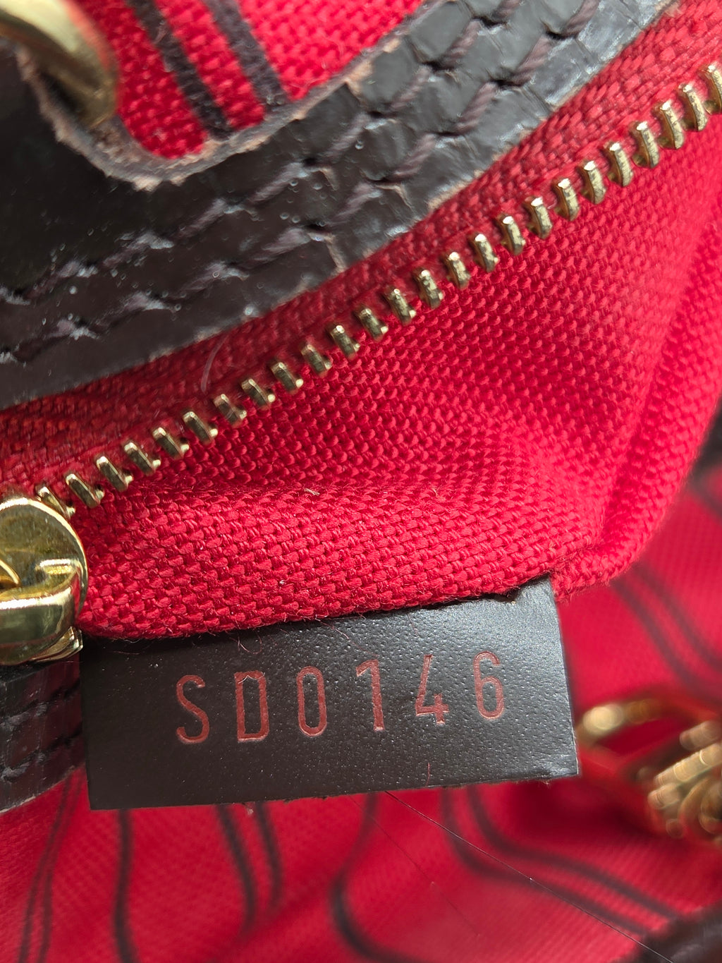 Close-up of a red woven fabric bag with a black leather tag stamped with the number 146DD0146, a gold zipper, and a gold metal pull.