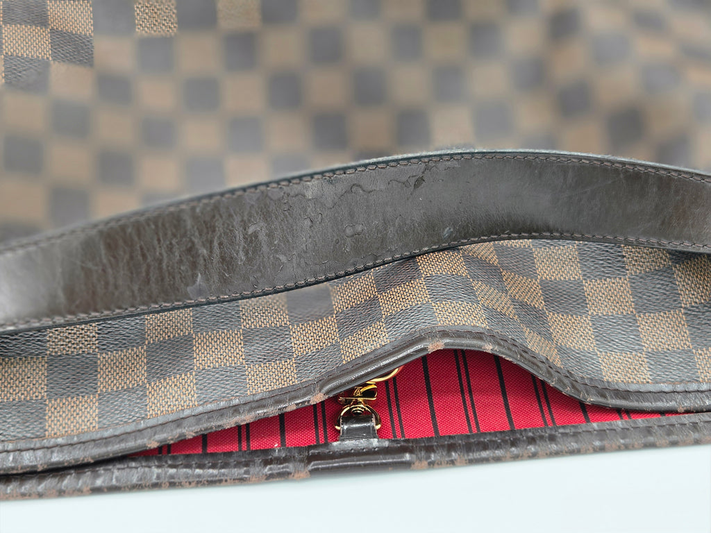 Close-up of a Louis Vuitton Damier canvas bag showing the interior lining, a worn dark brown leather trim, and a red striped inner flap.