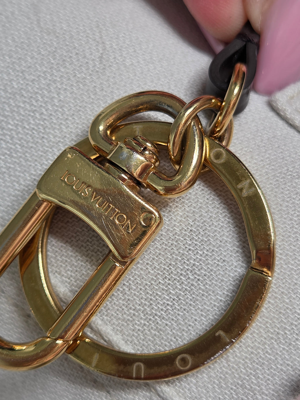 Close-up of a Louis Vuitton gold-tone metal key ring and clasp with a black strap, shown against a light fabric background.