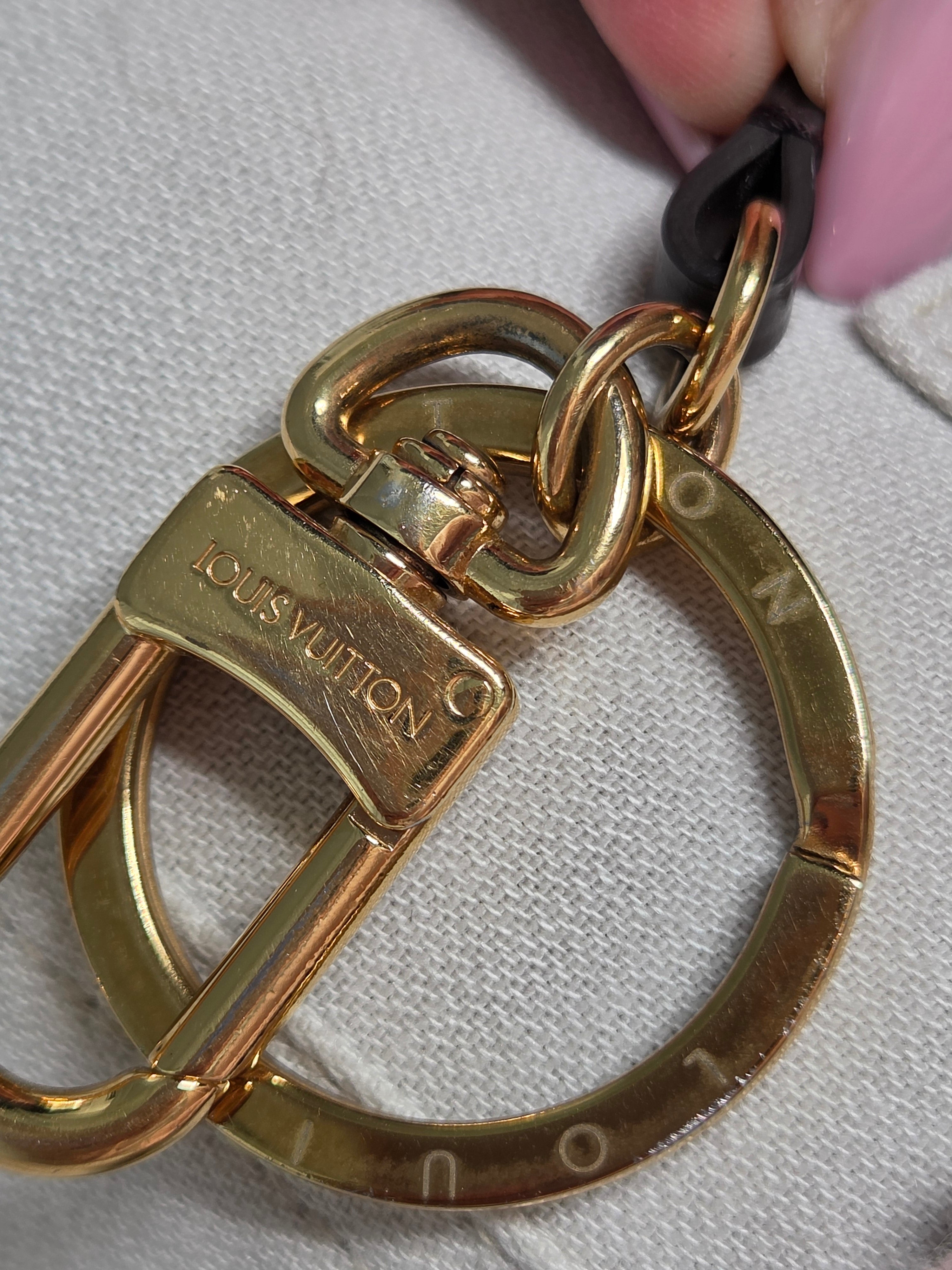 Close-up of a Louis Vuitton gold-tone metal key ring and clasp with a black strap, shown against a light fabric background.