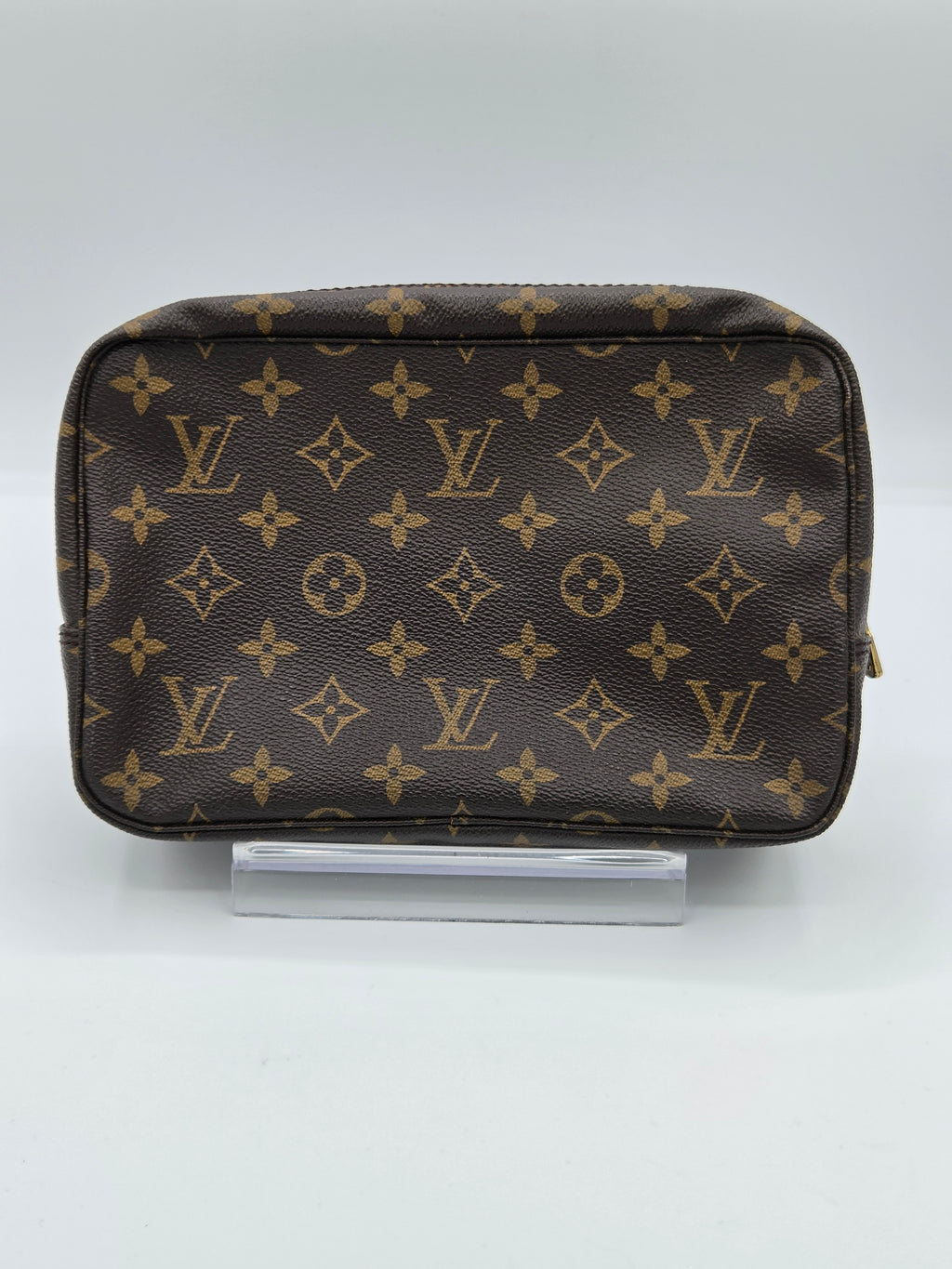 A Louis Vuitton monogram toiletry bag in brown coated canvas with gold-tone hardware, displayed on a clear acrylic stand against a light gray background.