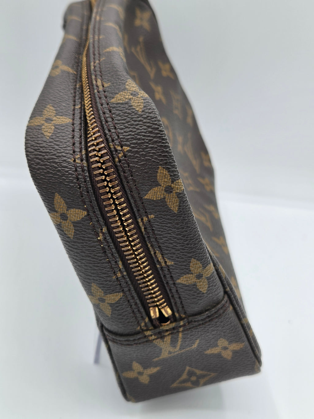 A close-up side view of a Louis Vuitton monogram canvas pouch with a gold zipper, showing the dark brown textured material and tan monogram pattern.