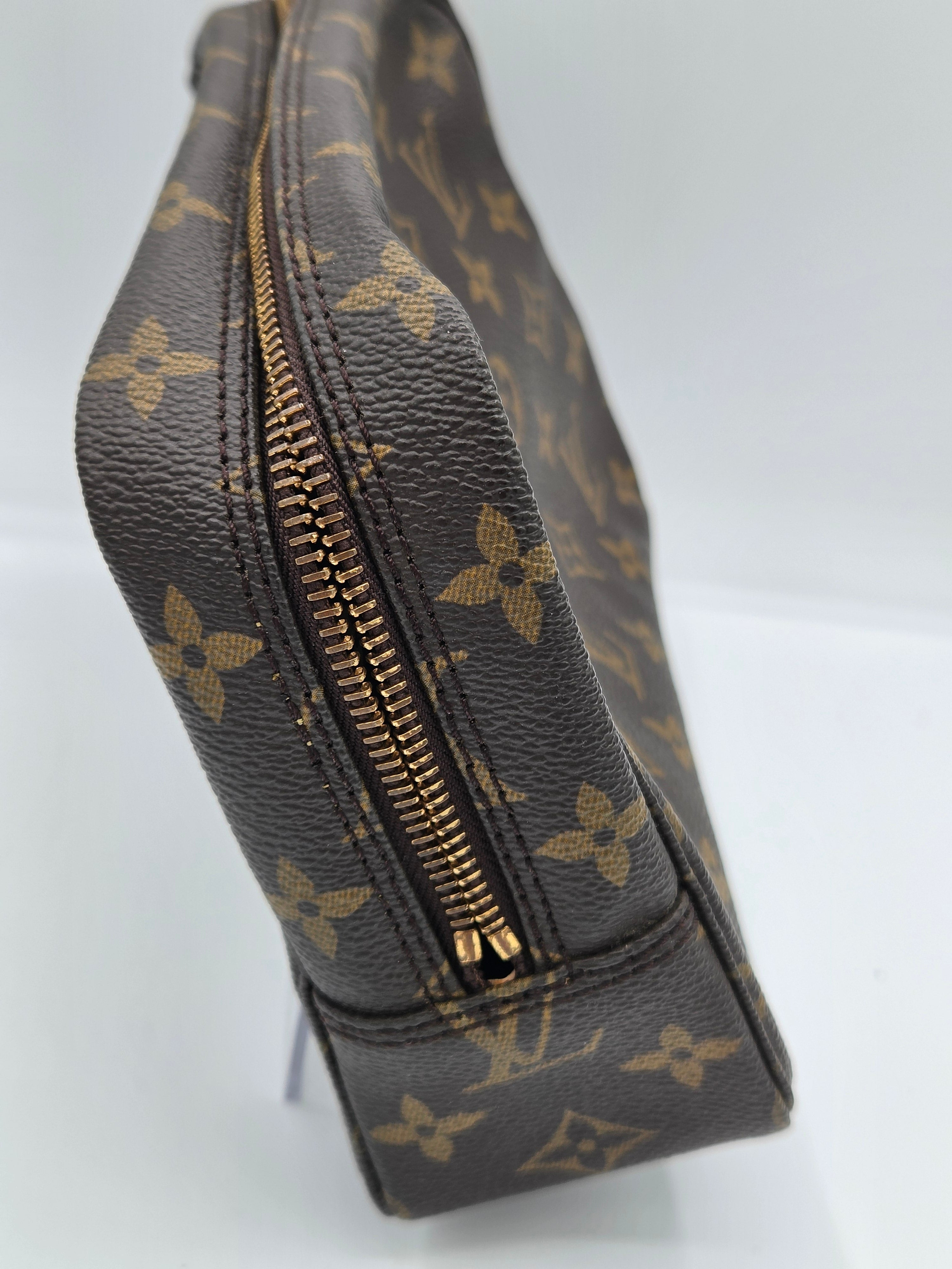 A close-up side view of a Louis Vuitton monogram canvas pouch with a gold zipper, showing the dark brown textured material and tan monogram pattern.