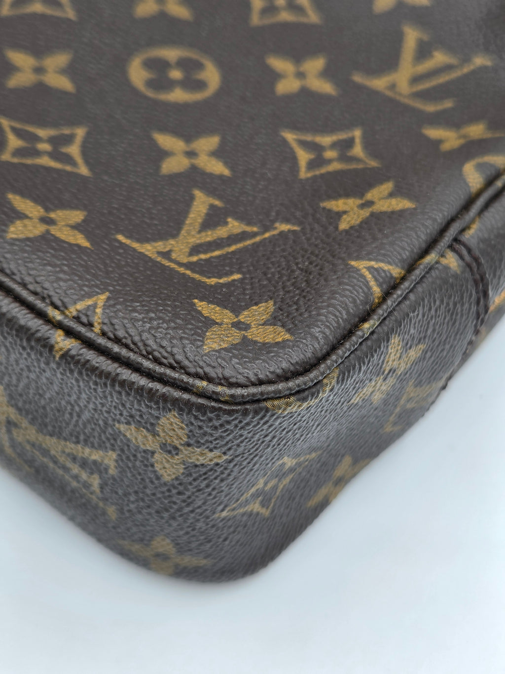 Close-up of a Louis Vuitton monogram canvas bag corner, showing the classic brown and gold LV pattern and pebbled leather texture on a white background.