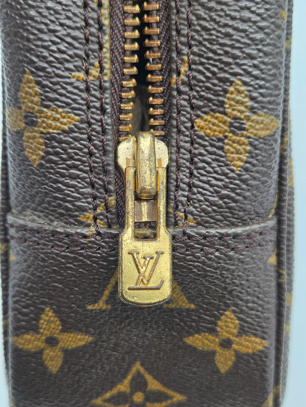 Close-up of a Louis Vuitton monogram canvas zipper with a gold-tone metal pull tab featuring the embossed LV logo.