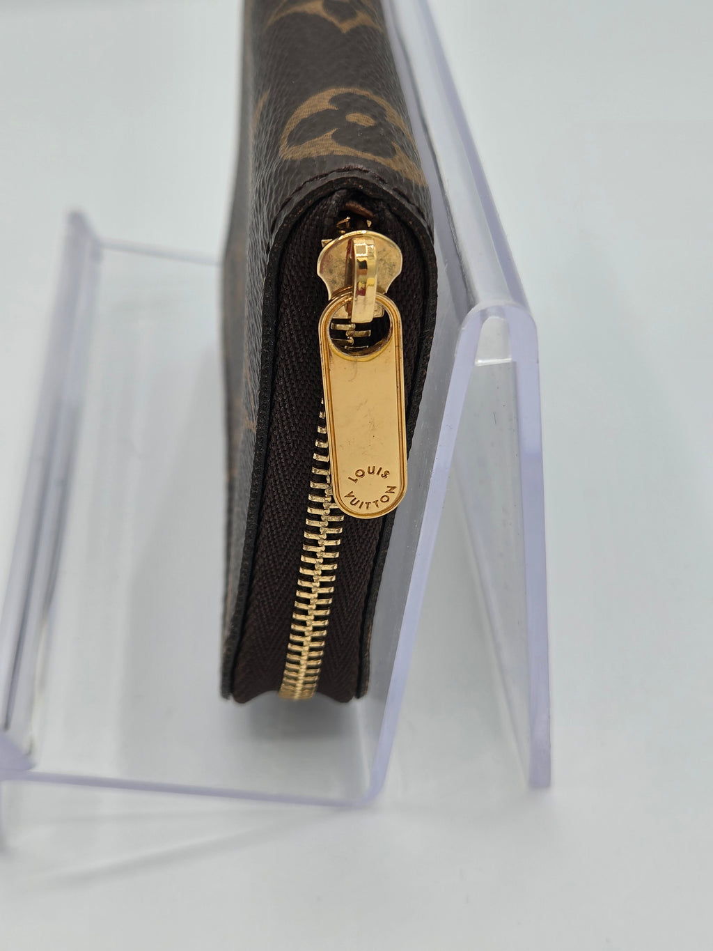 A close-up of a Louis Vuitton monogram wallet or zip case in dark brown coated canvas with gold-tone hardware, shown standing in a clear acrylic display stand on a white background.