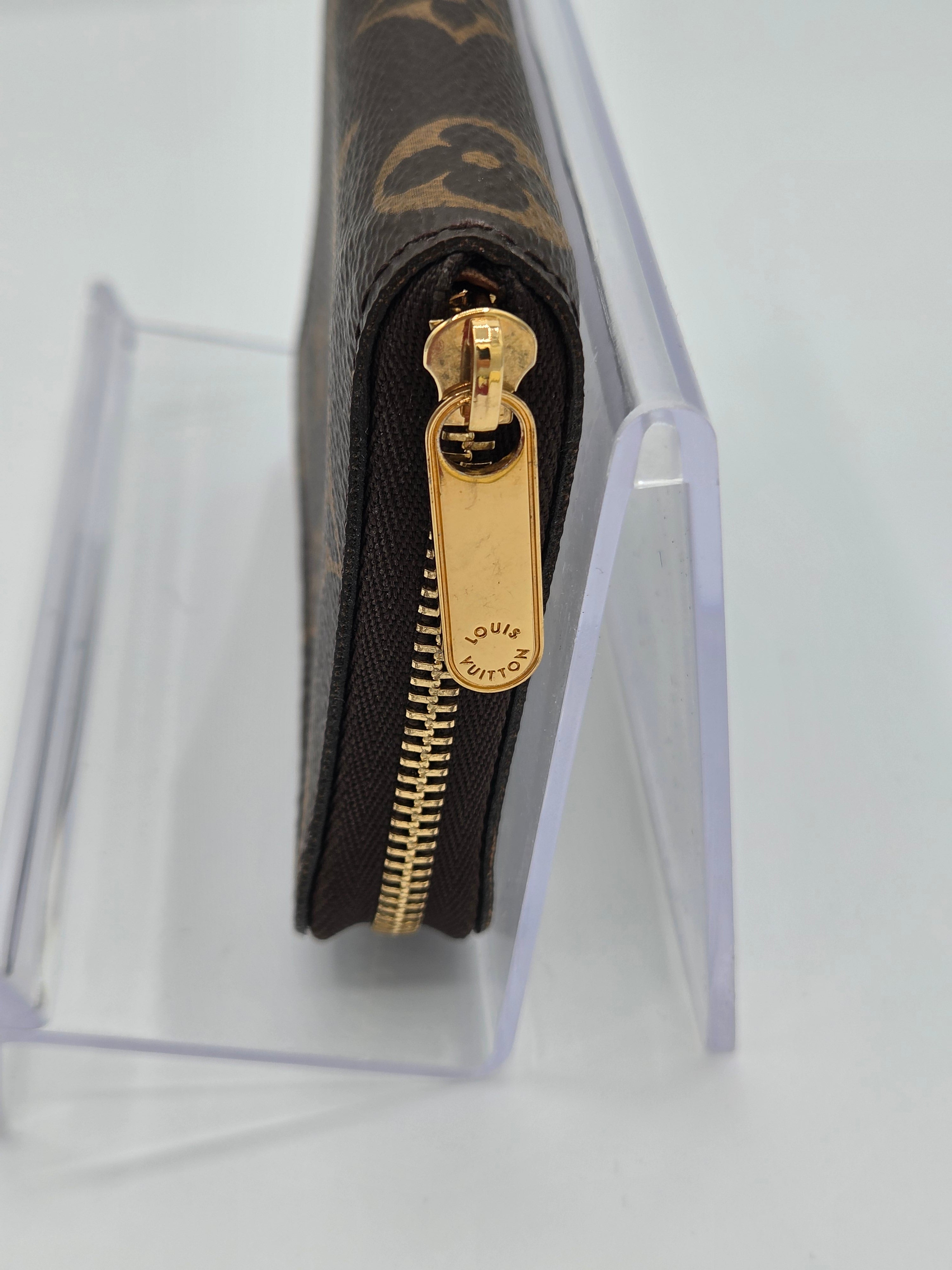 A close-up of a Louis Vuitton monogram wallet or zip case in dark brown coated canvas with gold-tone hardware, shown standing in a clear acrylic display stand on a white background.