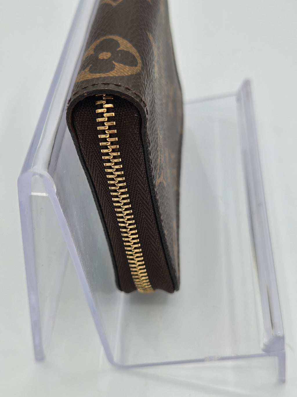 A close-up of a Louis Vuitton monogram wallet in brown coated canvas with a gold zipper, displayed on a clear acrylic stand against a white background.