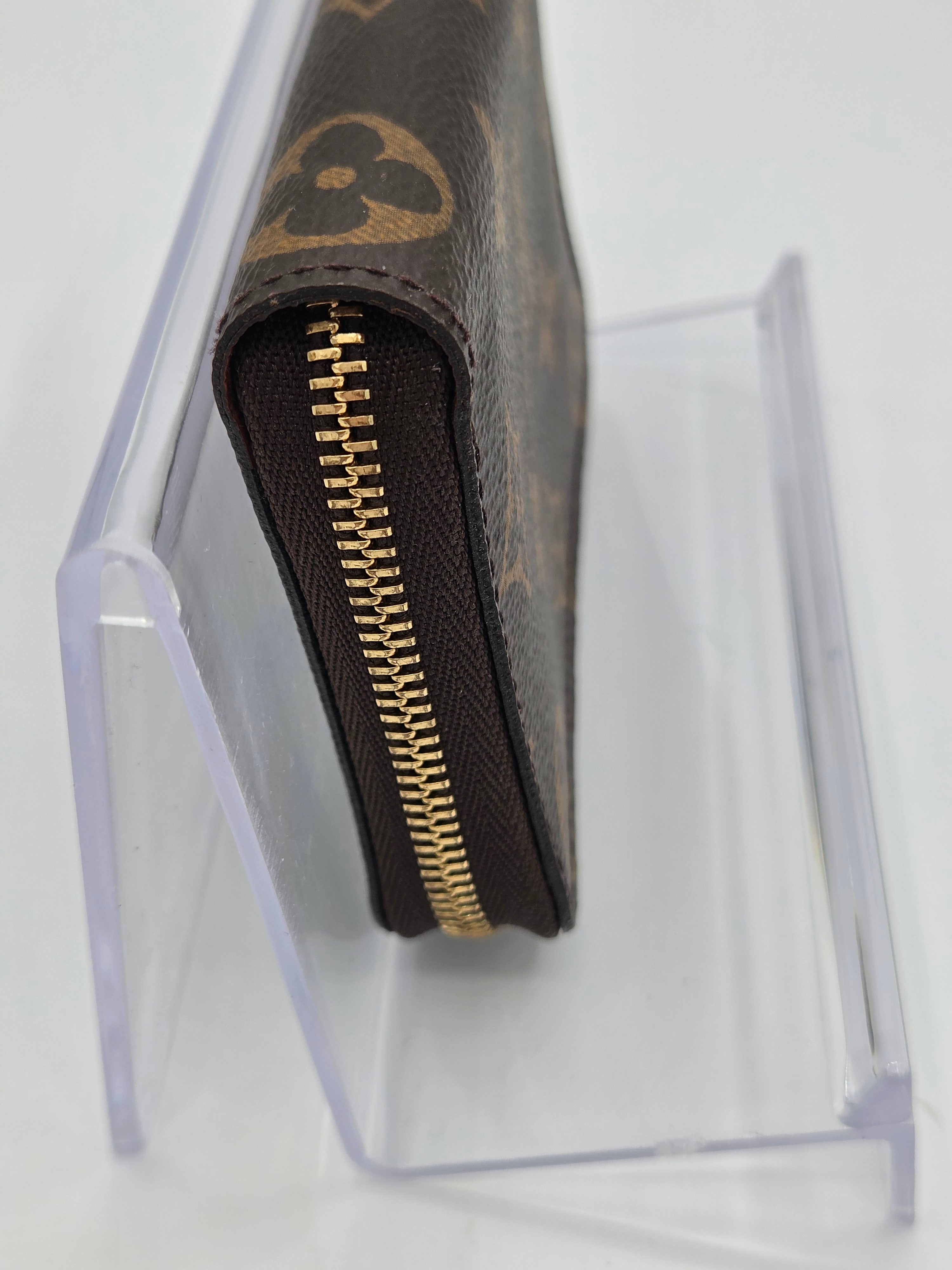 A close-up of a Louis Vuitton monogram wallet in brown coated canvas with a gold zipper, displayed on a clear acrylic stand against a white background.