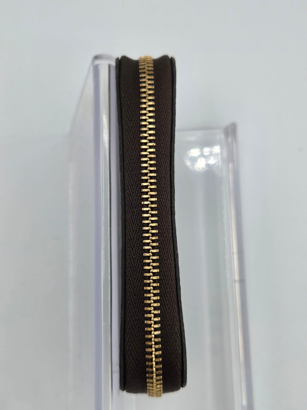 A close-up of a dark brown textured leather zipper pouch with a prominent gold-tone metal zipper, shown standing vertically in a clear plastic display stand against a white background.