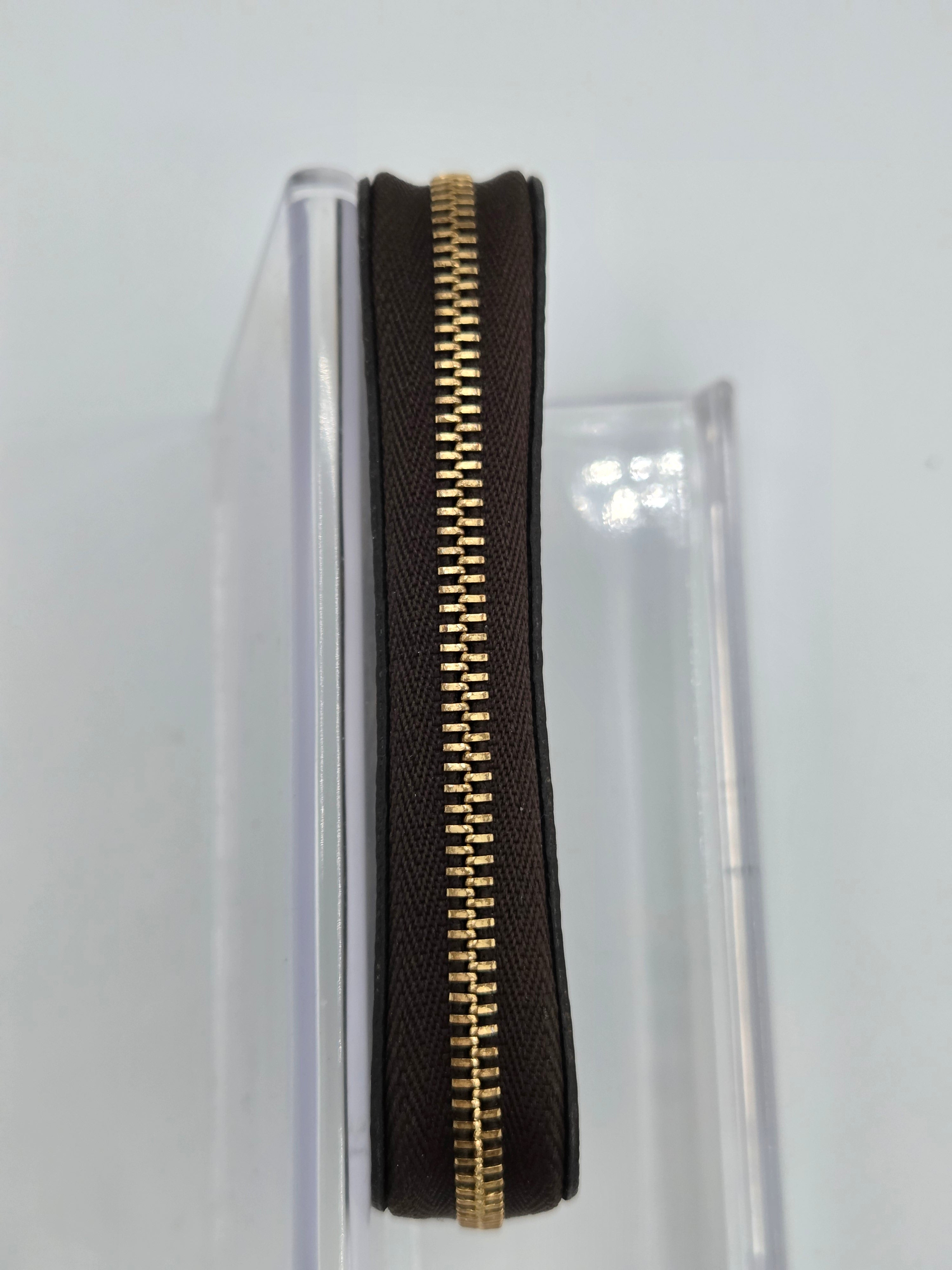 A close-up of a dark brown textured leather zipper pouch with a prominent gold-tone metal zipper, shown standing vertically in a clear plastic display stand against a white background.