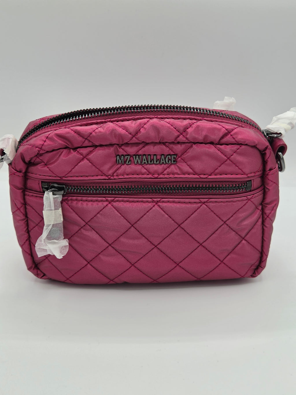 A pink quilted crossbody bag with black zippers and the brand name 'MZ WALLACE' printed on the front, shown on a light gray background with protective plastic still attached to the zipper pulls.