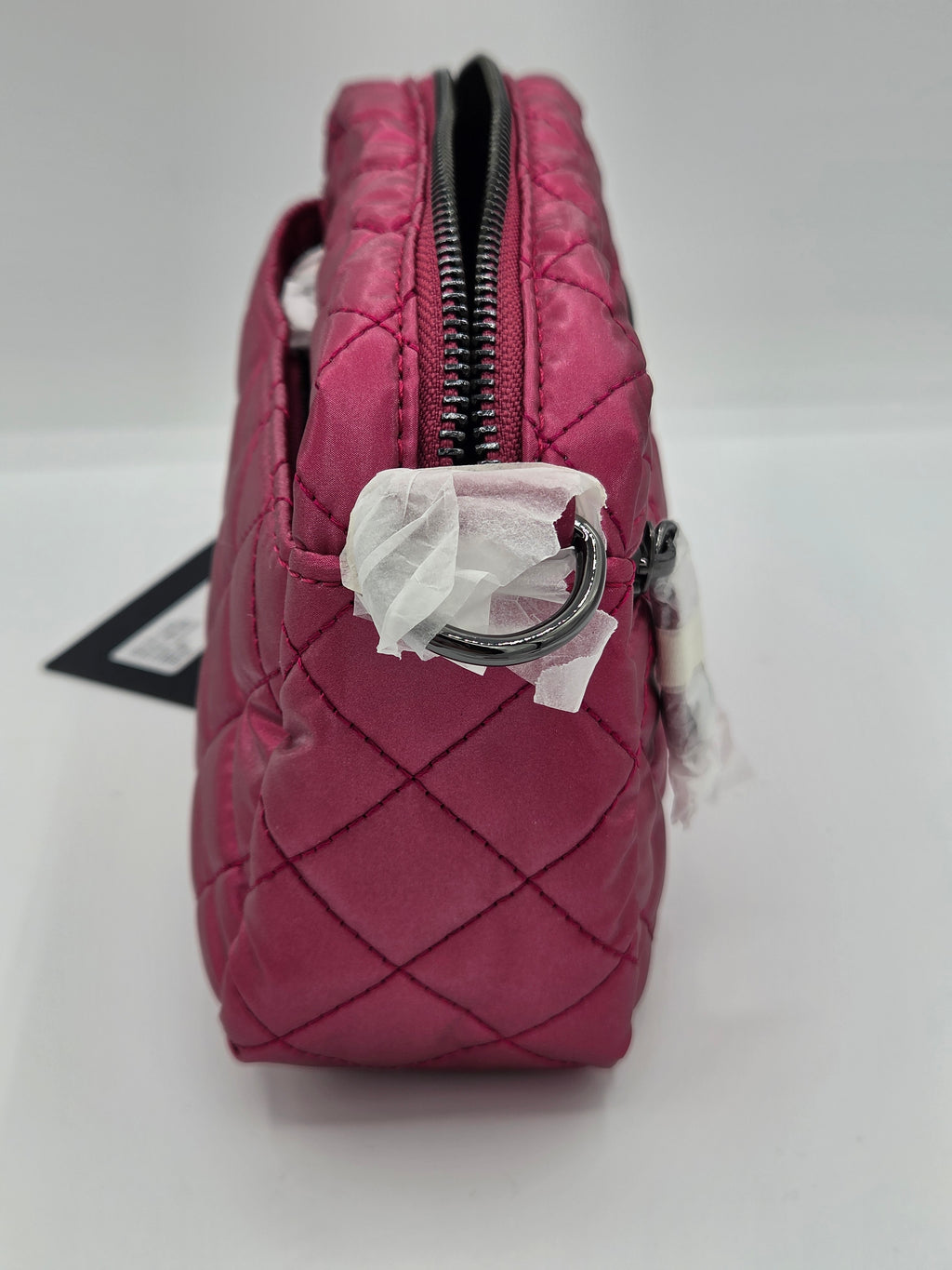 A close-up of a quilted pink handbag or pouch with a black zipper, metal D-ring hardware, and white tissue paper wrapping around the zipper, set against a plain white background.