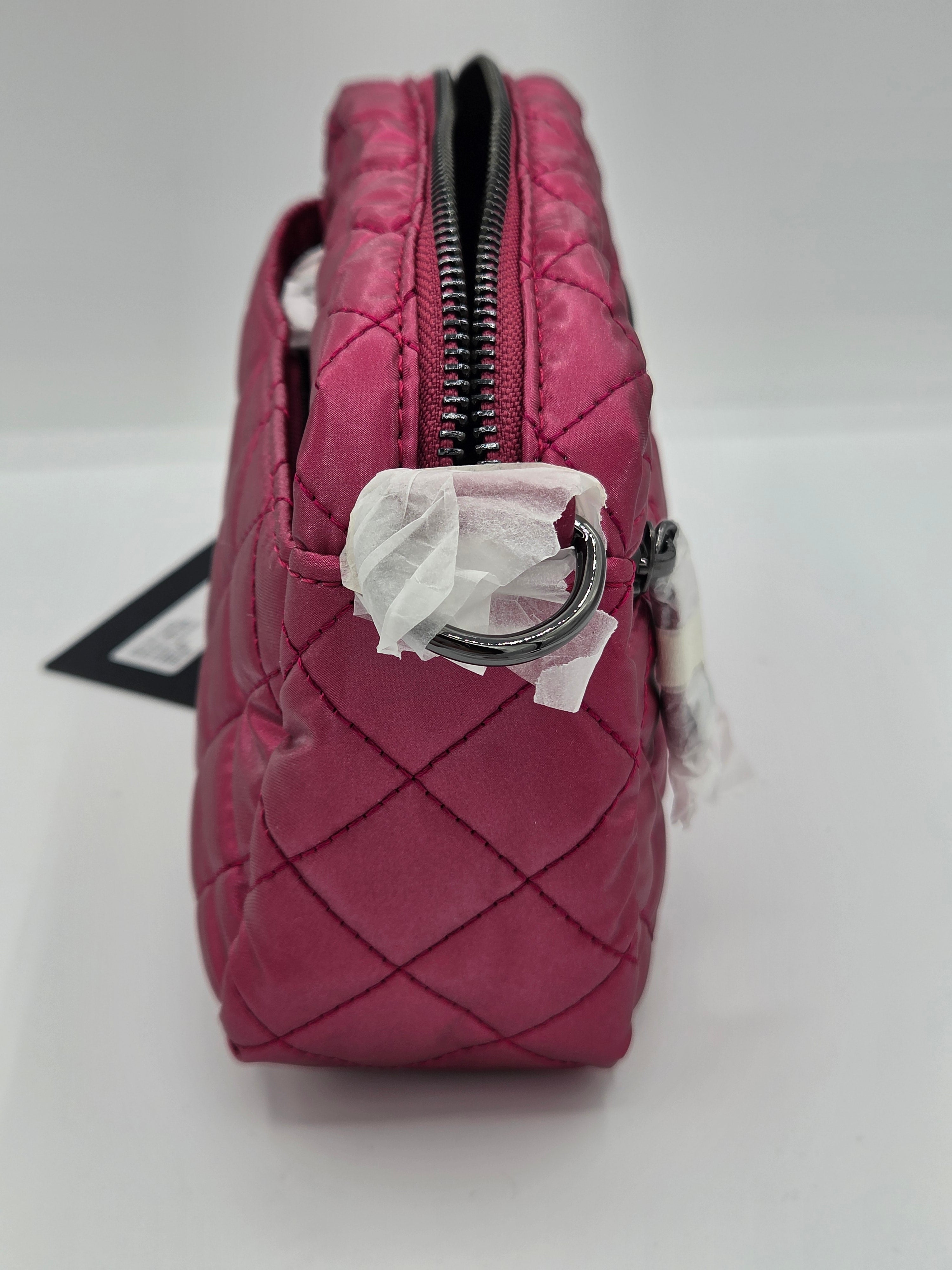 A close-up of a quilted pink handbag or pouch with a black zipper, metal D-ring hardware, and white tissue paper wrapping around the zipper, set against a plain white background.