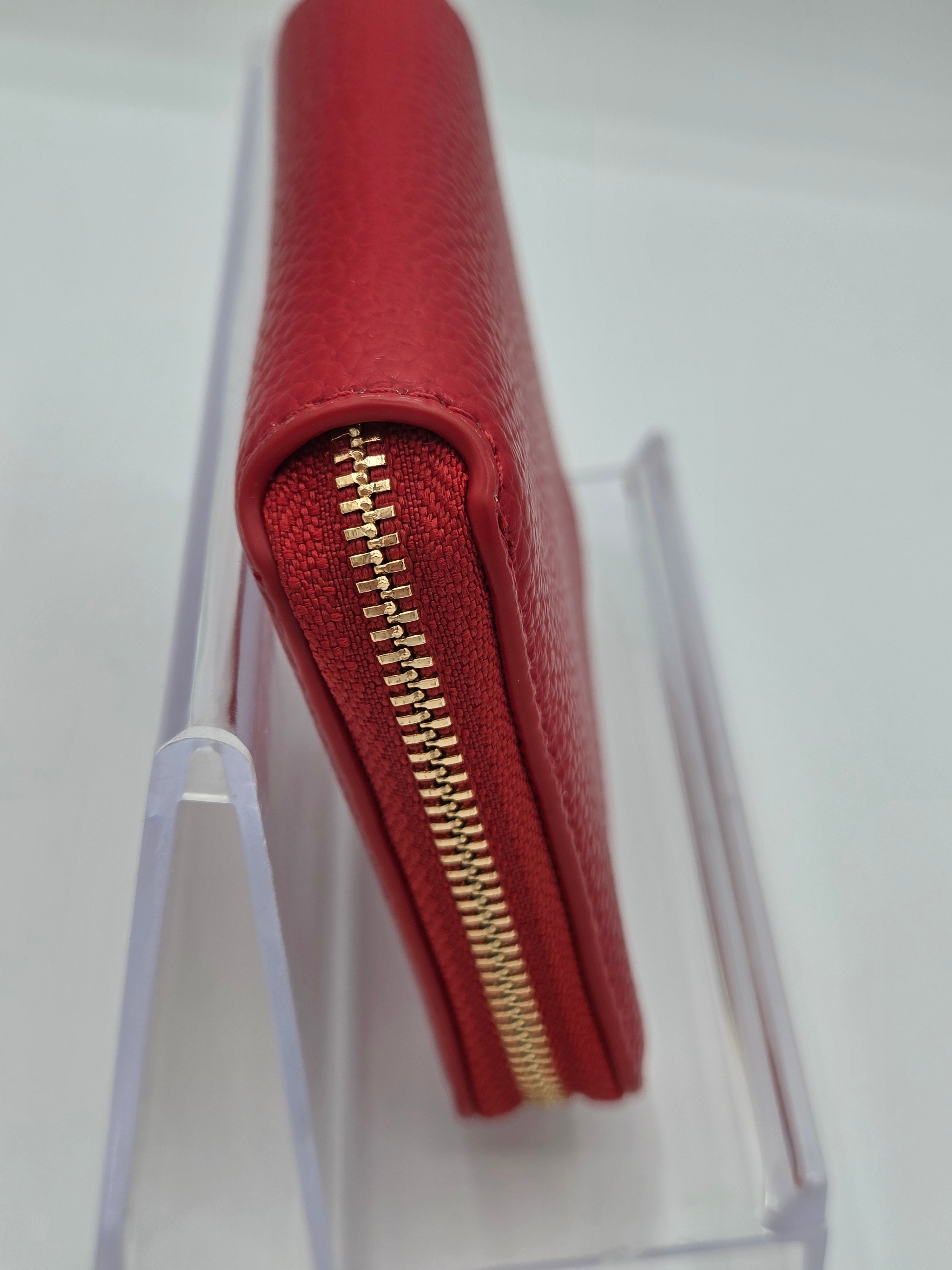 A red textured leather zip-around wallet standing upright in a clear acrylic display stand, showing a gold-tone zipper along the side.