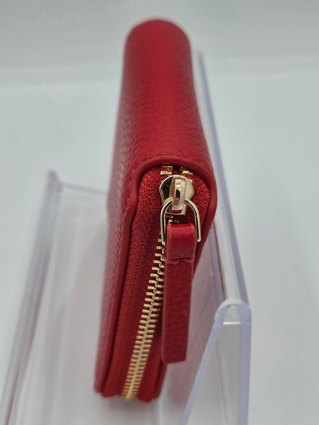A red pebbled leather zip-around wallet with gold-tone hardware, shown standing upright in a clear acrylic display stand against a white background.