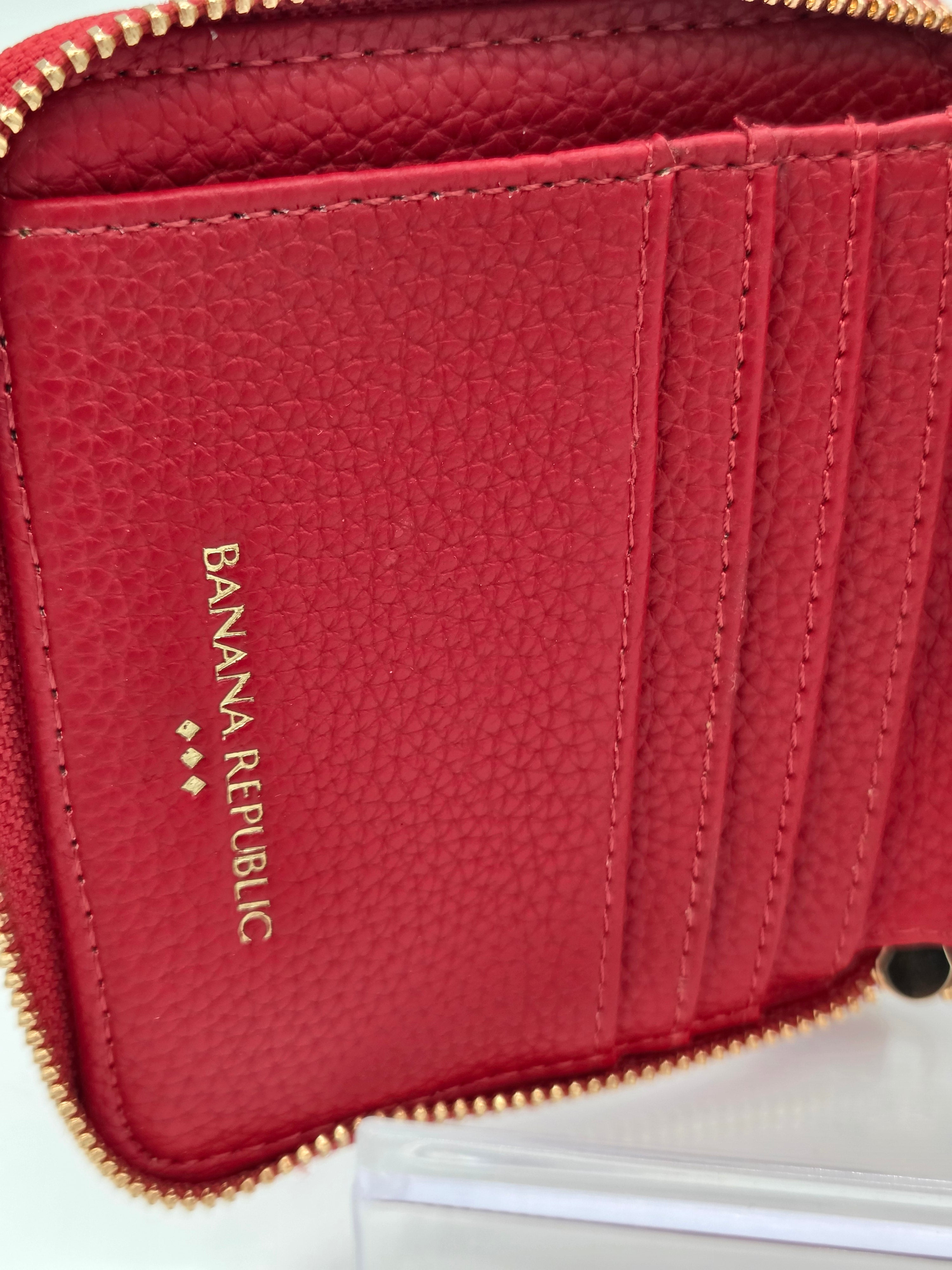 Close-up of a red pebbled leather zip-around wallet with gold-tone hardware and vertical card slots. The brand name 'BANANA REPUBLIC' is printed vertically in gold on the inside pocket.