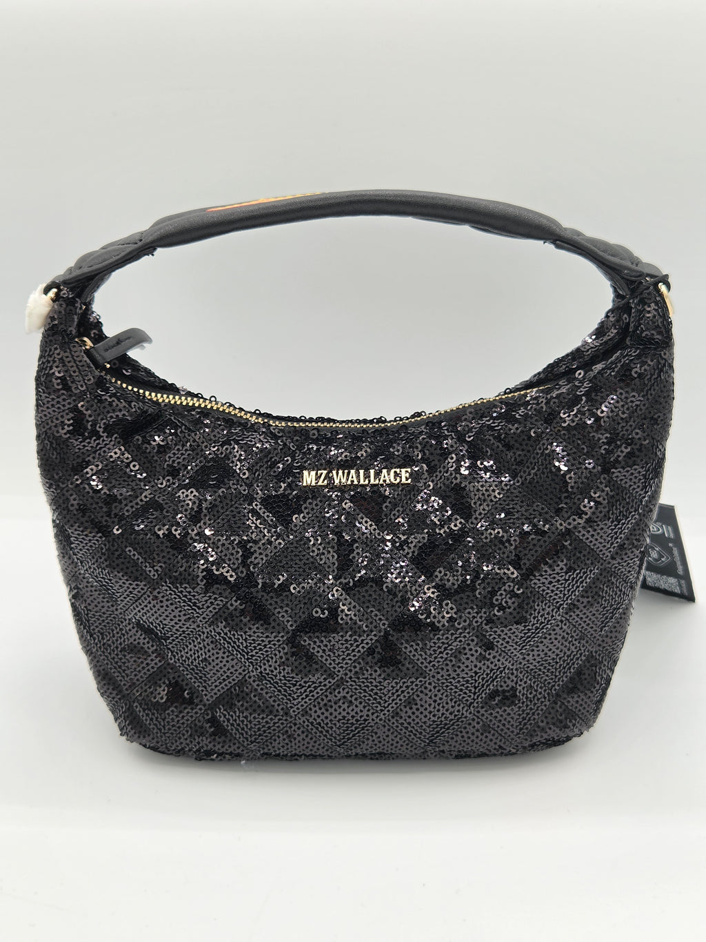 A black sequined hobo handbag with a gold-tone 'MZ WALLACE' logo plaque on the front, a black leather handle, and a small black hangtag attached on the right side, set against a plain light gray background.