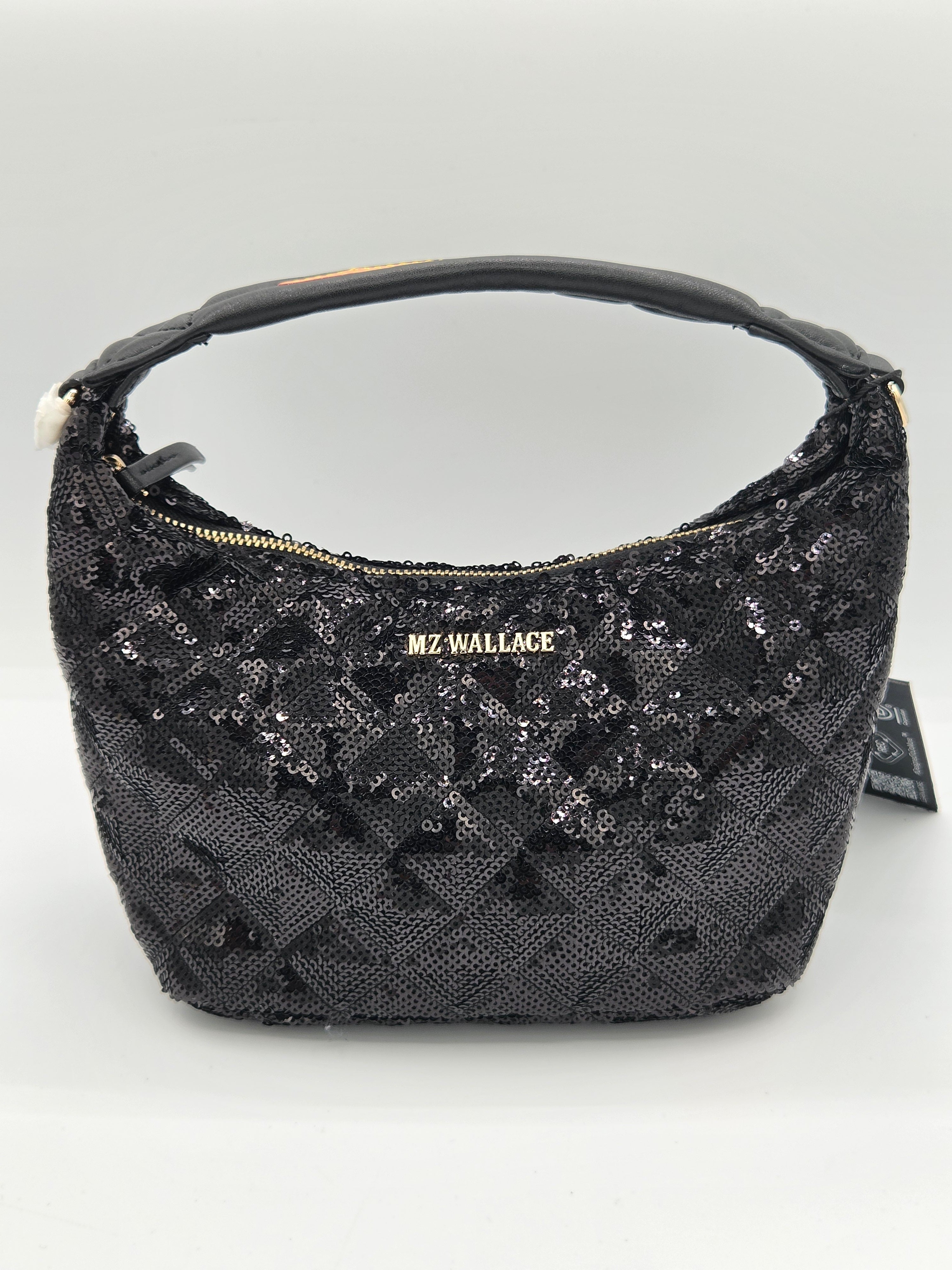 A black sequined hobo handbag with a gold-tone 'MZ WALLACE' logo plaque on the front, a black leather handle, and a small black hangtag attached on the right side, set against a plain light gray background.