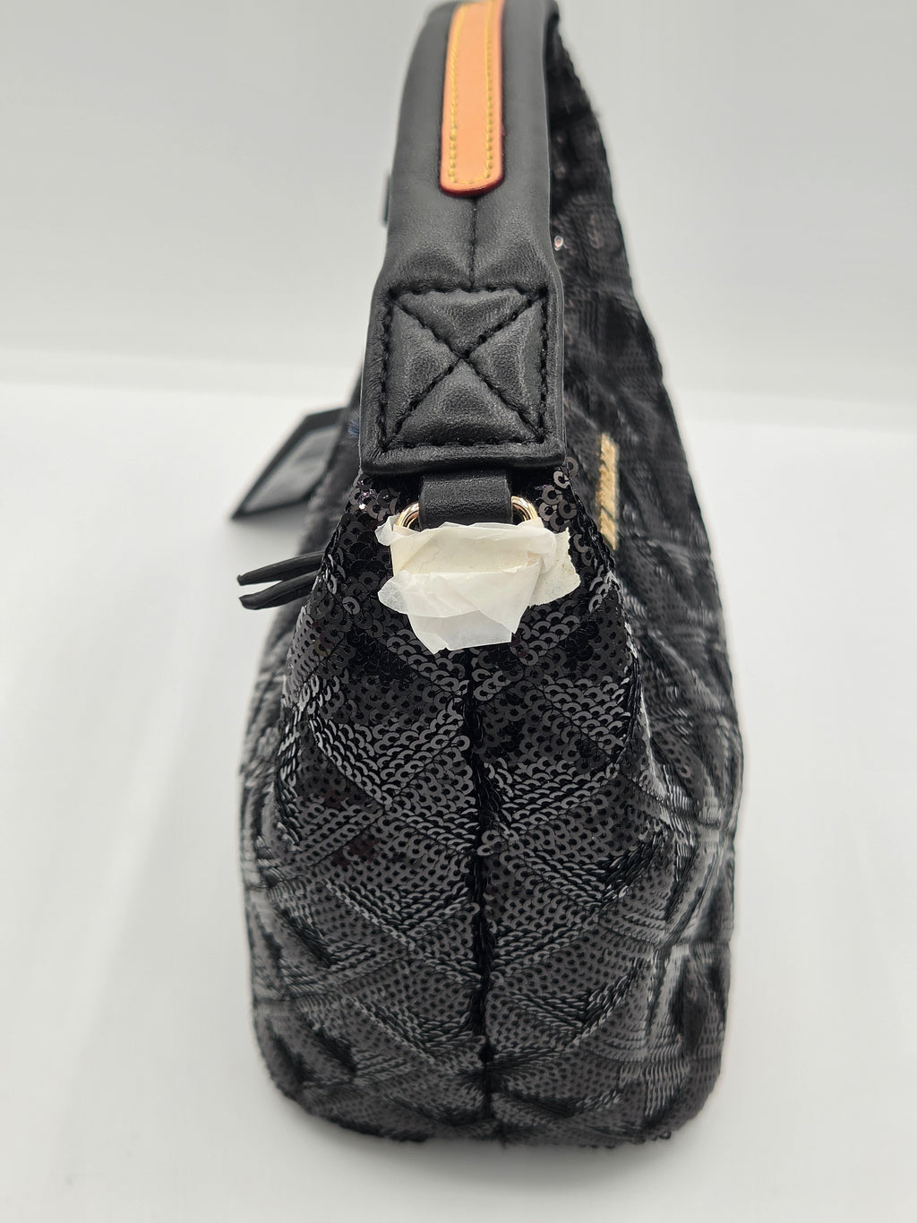 A close-up side view of a black quilted handbag with a textured woven pattern, featuring a black leather strap with an orange accent tab and a white paper protective insert at the zipper.