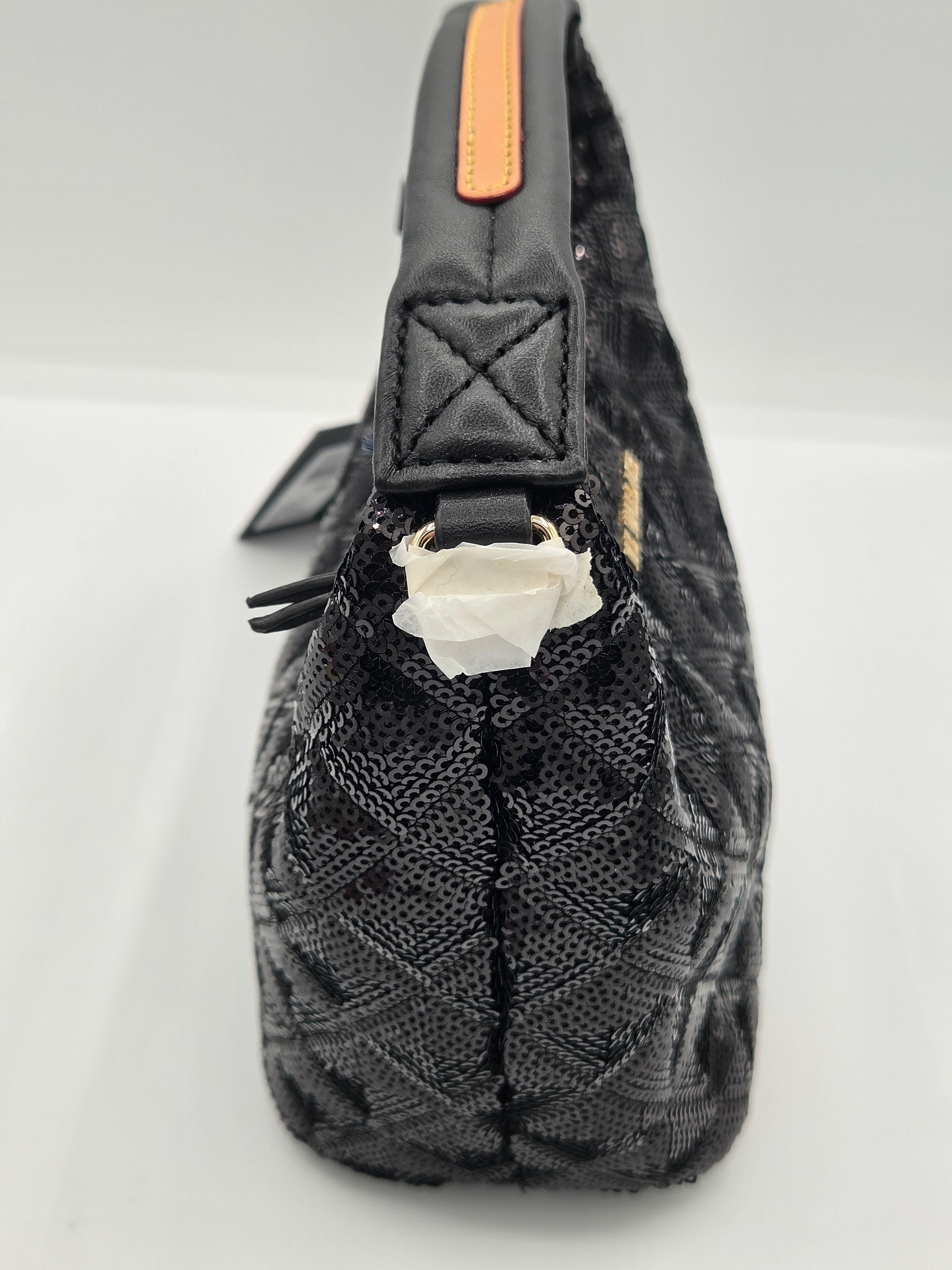 A close-up side view of a black quilted handbag with a textured woven pattern, featuring a black leather strap with an orange accent tab and a white paper protective insert at the zipper.