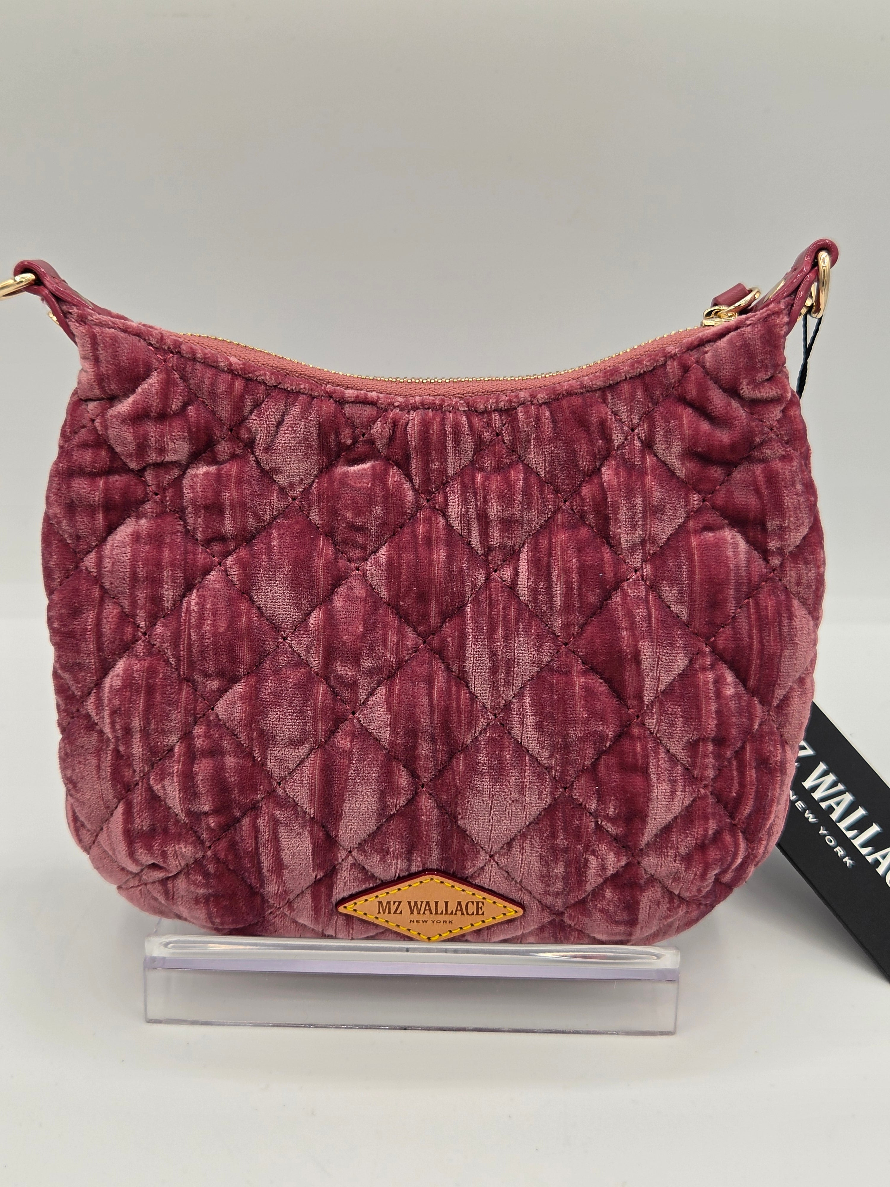 A quilted plum velvet shoulder bag with gold-tone hardware, displayed on a clear acrylic stand with its black brand tag visible on the right.