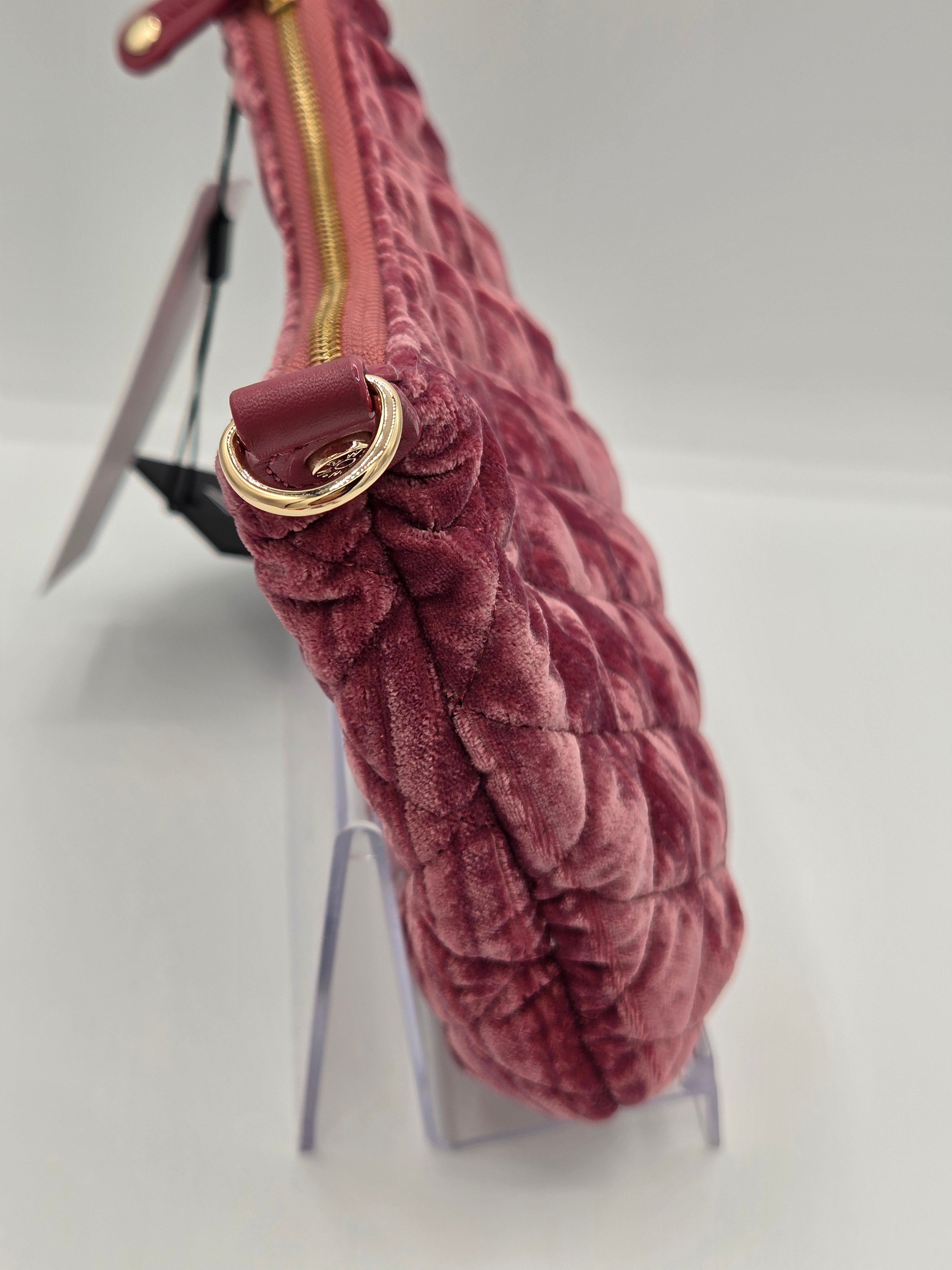 A close-up of a dusty rose velvet clutch bag with a gold-tone zipper and a gold-tone D-ring, displayed on a clear acrylic stand.