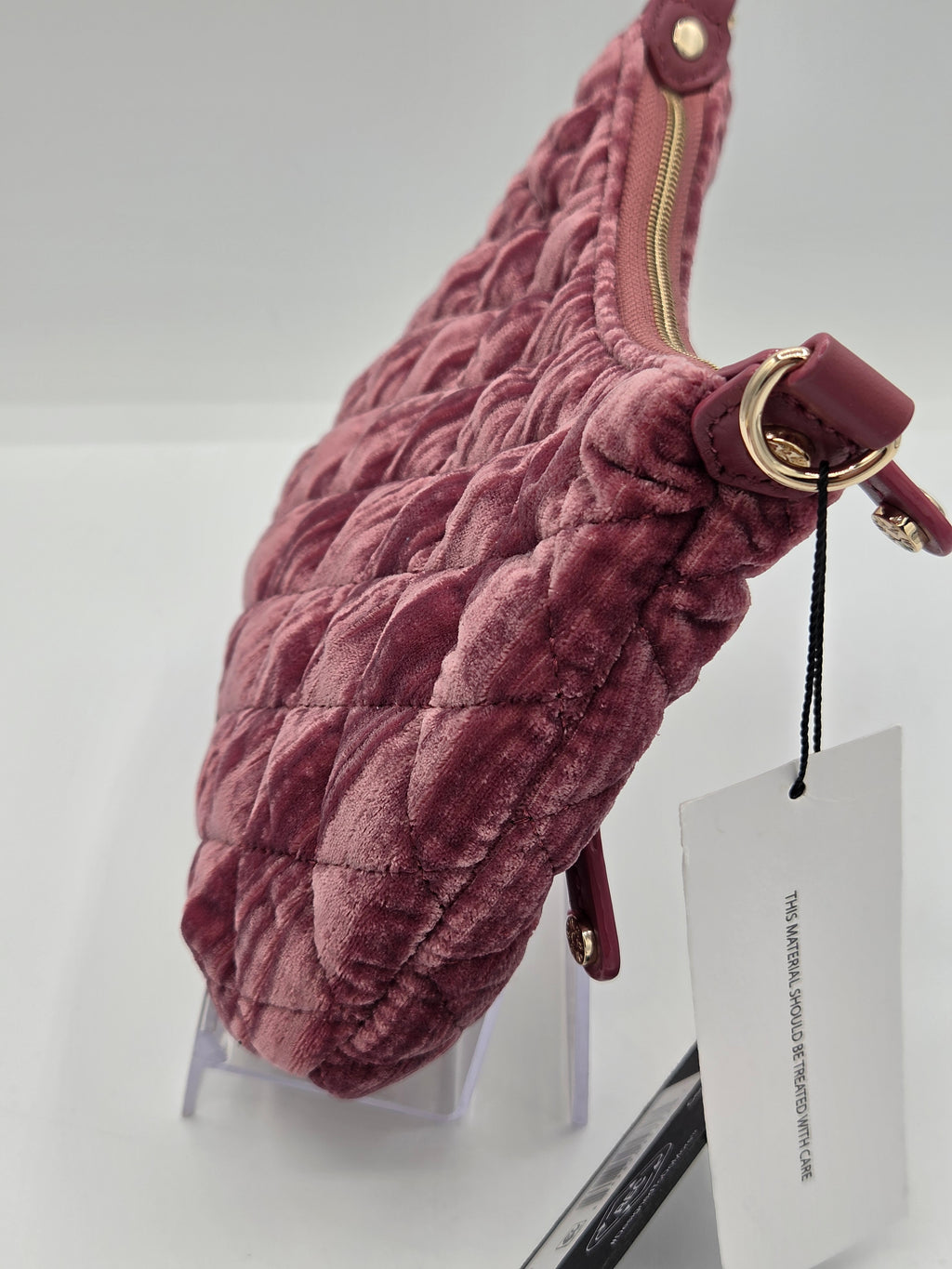 A dusty rose quilted velvet shoulder bag with gold-tone hardware, displayed on a clear stand with a white product tag attached.