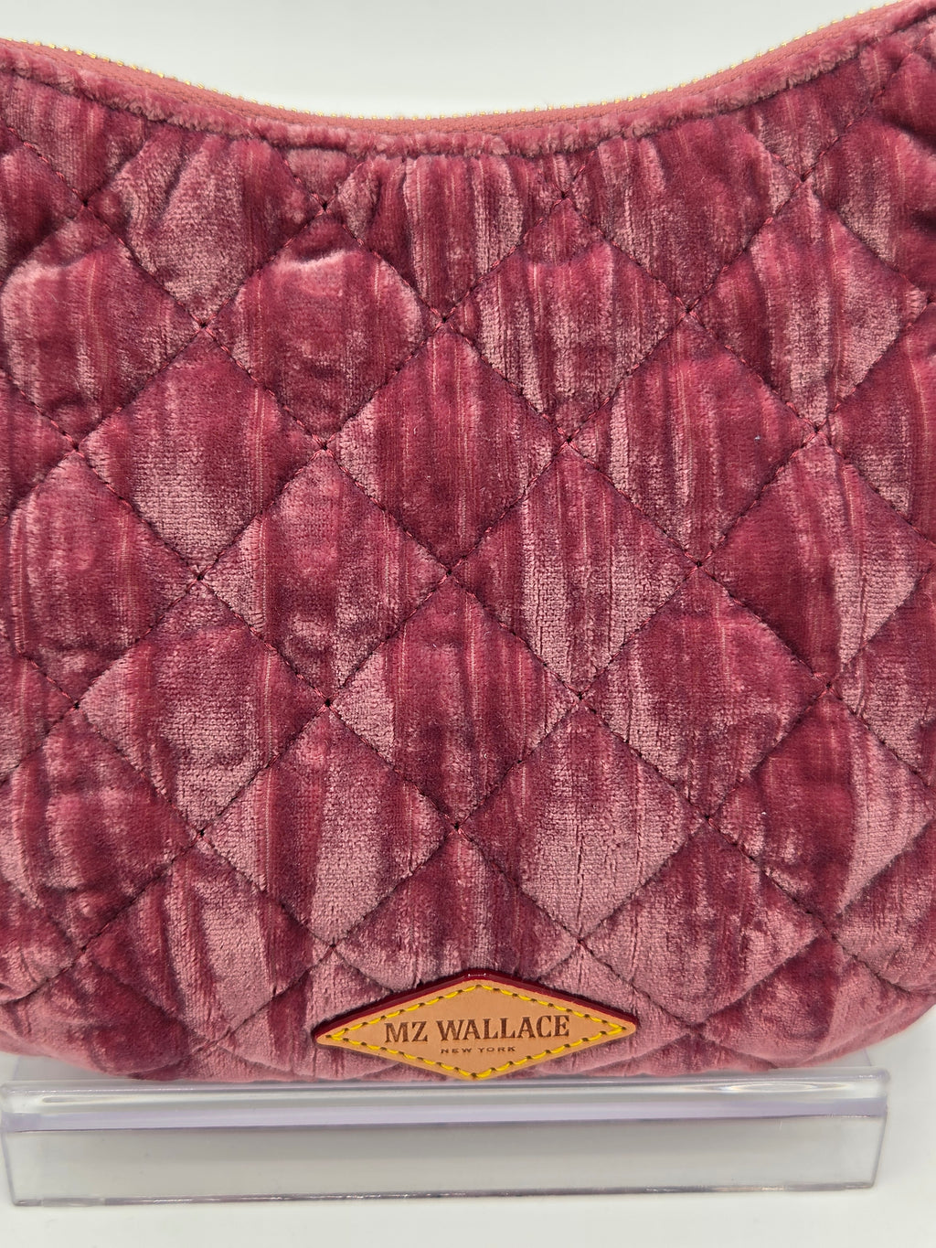 A close-up of a quilted dusty rose velvet pouch bag with a diamond pattern and a yellow diamond-shaped 'MZ WALLACE NEW YORK' logo patch on the front, resting on a clear acrylic stand.