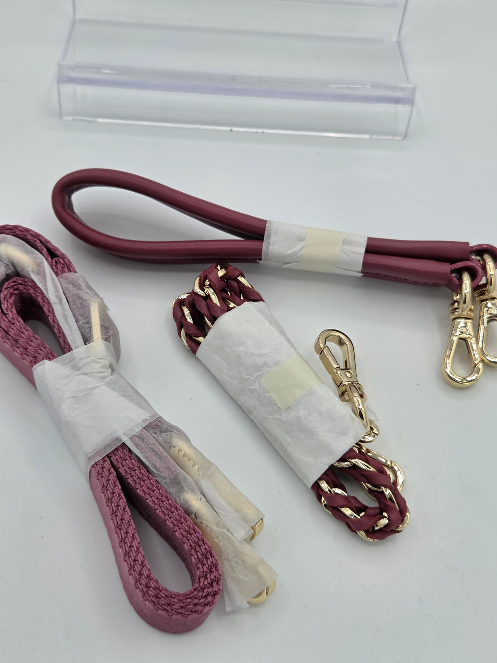 Two leather and braided strap accessories for a bag, wrapped in protective paper and clear plastic. One strap is a smooth burgundy leather loop with gold-tone hardware, and the other is a burgundy leather and gold-tone braided rope strap.