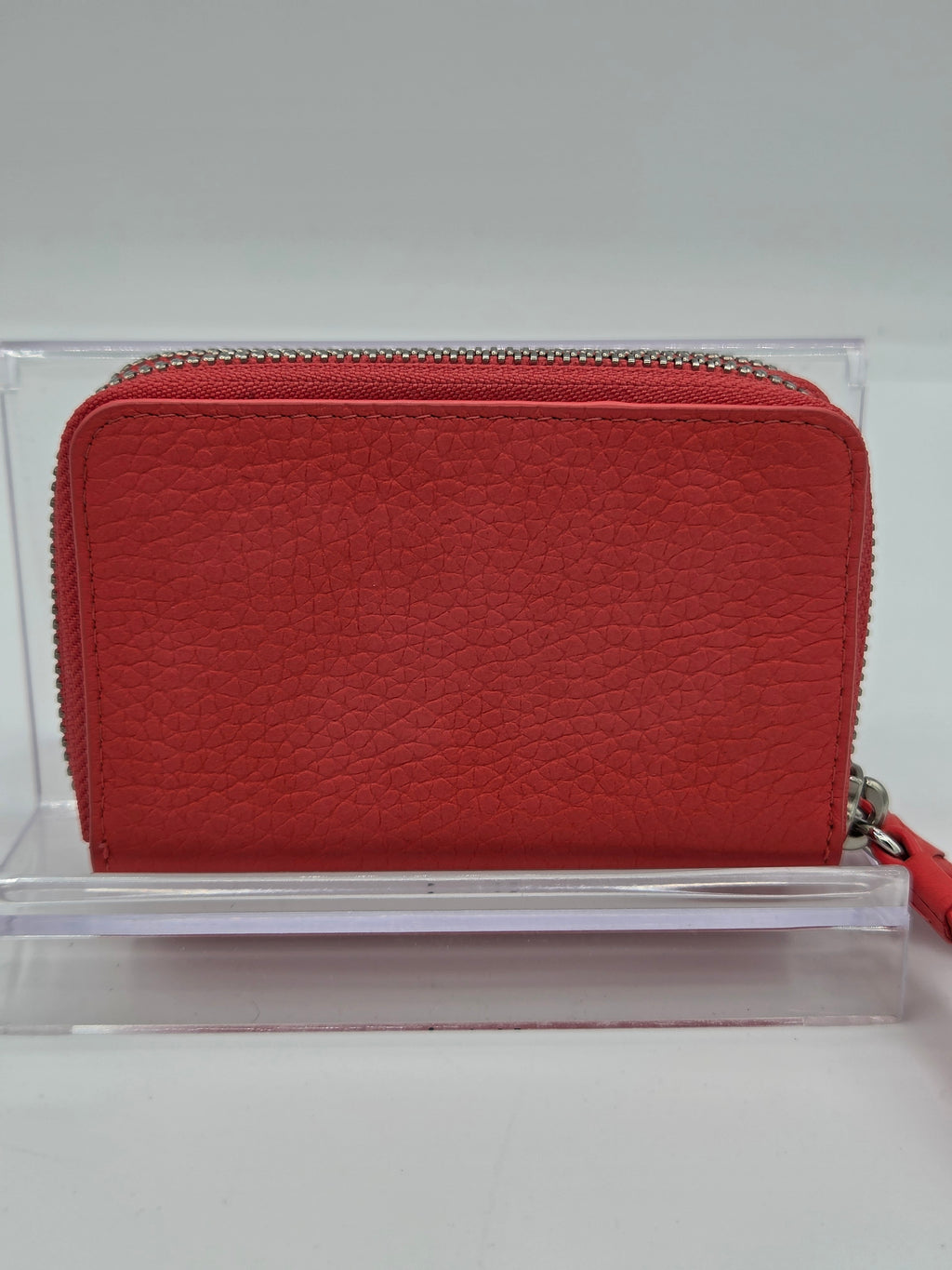 A red pebbled leather zip-around wallet with a silver-tone zipper, displayed on a clear acrylic stand against a light gray background.
