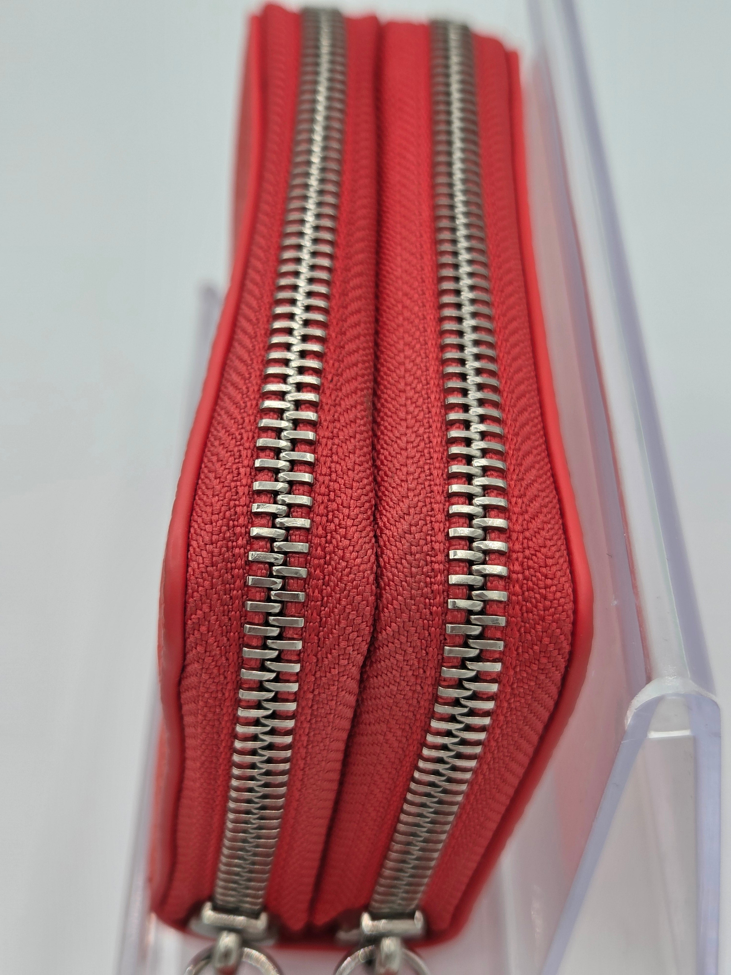 A close-up of a red zip-around wallet or coin purse with two silver metal zippers, shown inside a clear plastic display box.