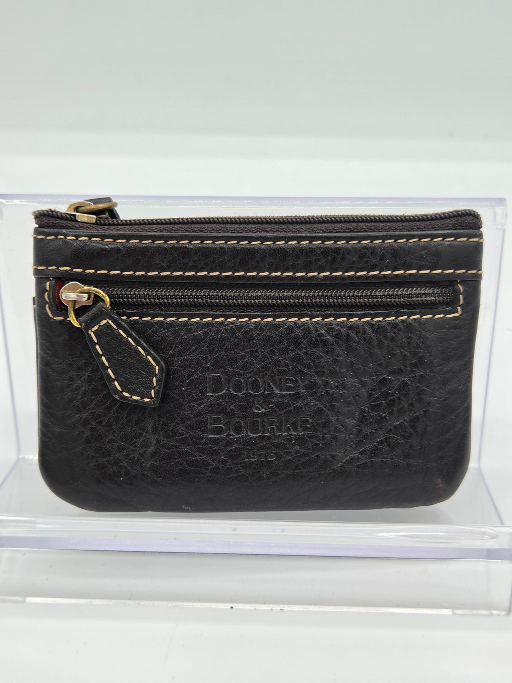 A black textured leather coin pouch with gold-toned hardware and contrast stitching, featuring an embossed 'DOONEY & BOURGA' logo, displayed inside a clear acrylic case.