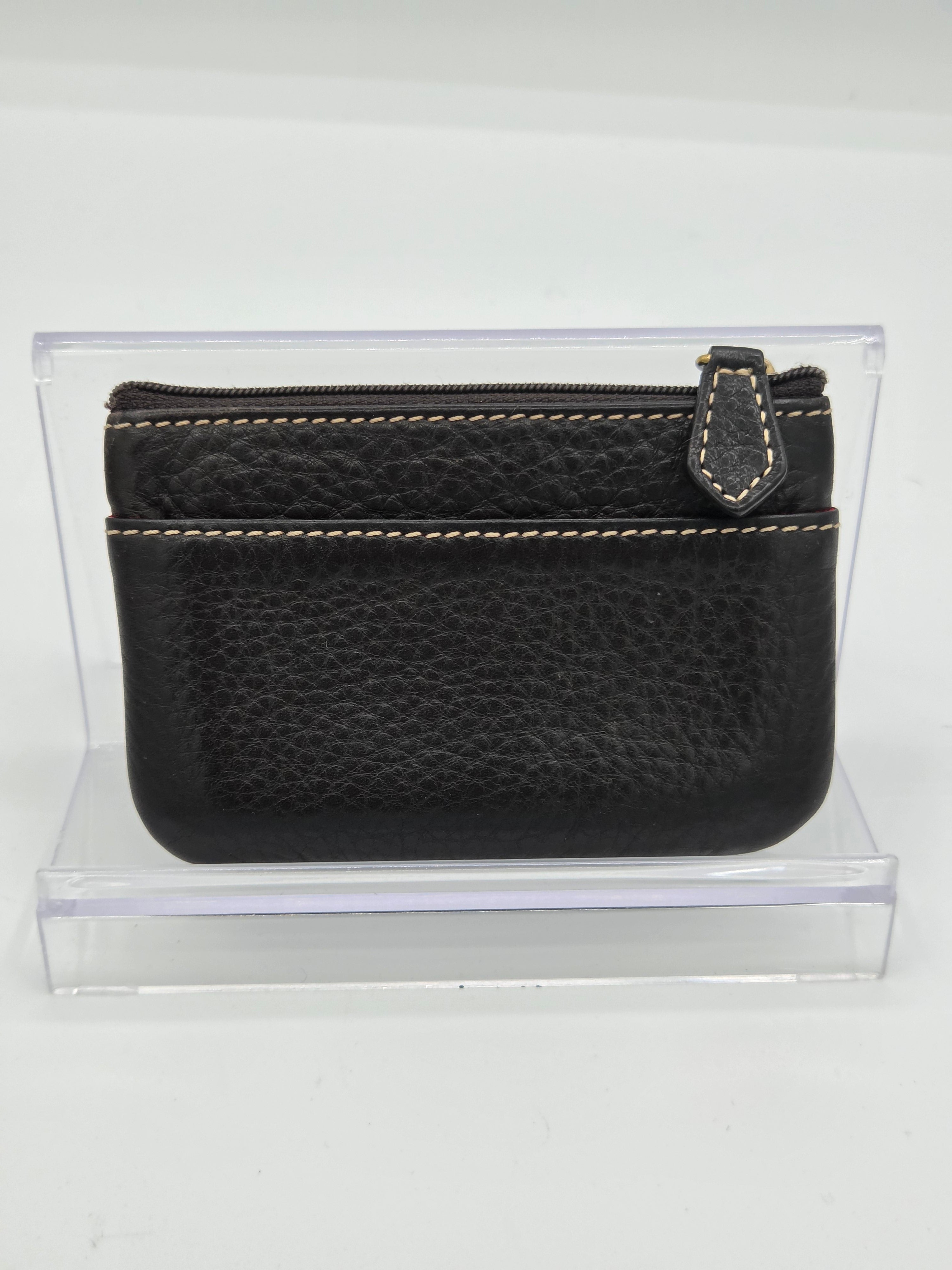 A black pebbled leather zip-around pouch with light tan contrast stitching, displayed inside a clear acrylic stand against a white background.