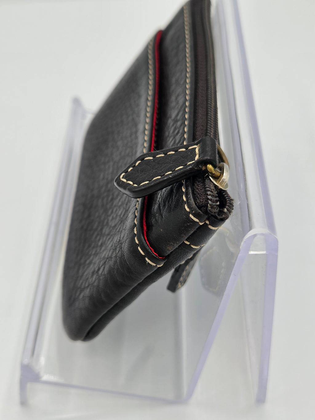 A black pebbled leather zip-around clutch wallet with cream contrast stitching and a red accent line, displayed on a clear acrylic stand.