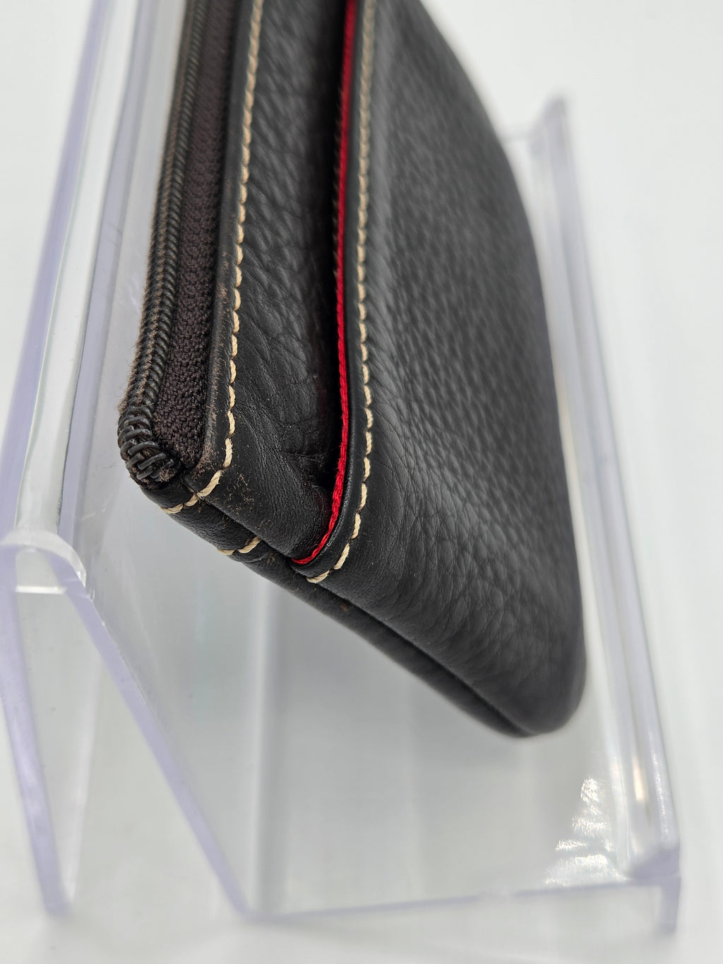 A close-up of a black textured leather zippered pouch or wallet with contrast stitching and a red accent line, displayed on a clear acrylic stand against a white background.