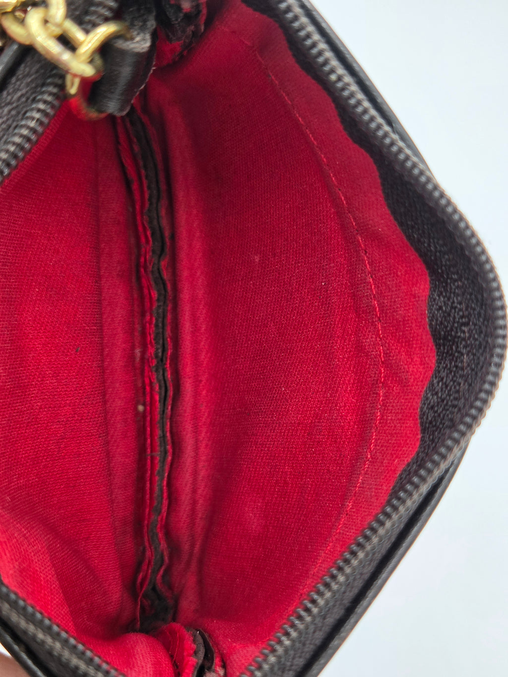 Close-up of the interior of a black textured handbag or wallet, showing a bright red fabric lining with a central zipper and a gold-tone metal pull at the top.