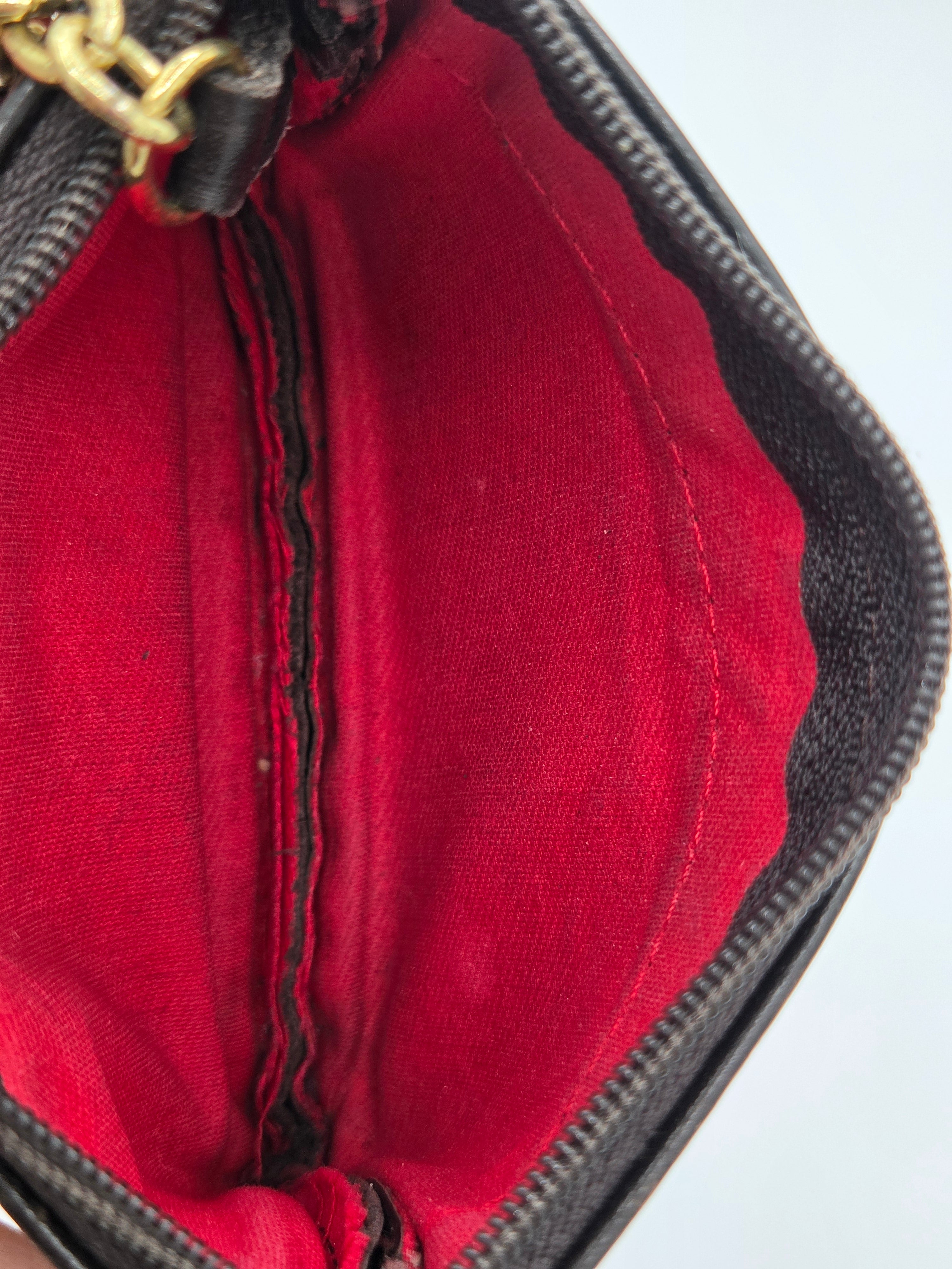 Close-up of the interior of a black textured handbag or wallet, showing a bright red fabric lining with a central zipper and a gold-tone metal pull at the top.