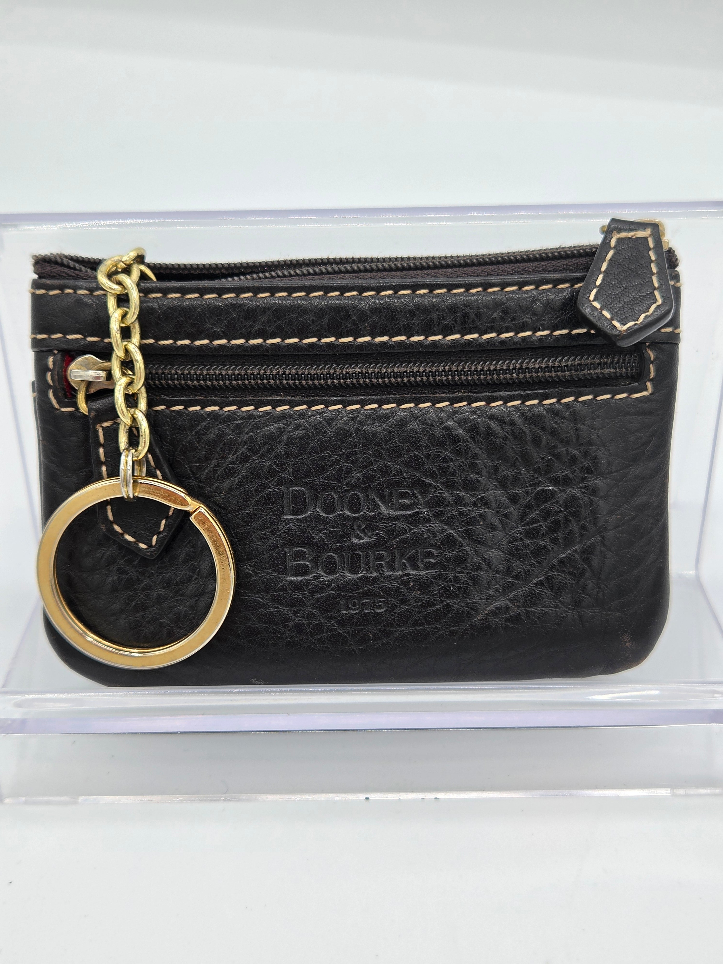 A black textured leather coin purse with gold-tone hardware, featuring a gold chain and key ring, double zipper compartments, and an embossed 'Dooney & Bourke' logo on the front, displayed inside a clear plastic case.