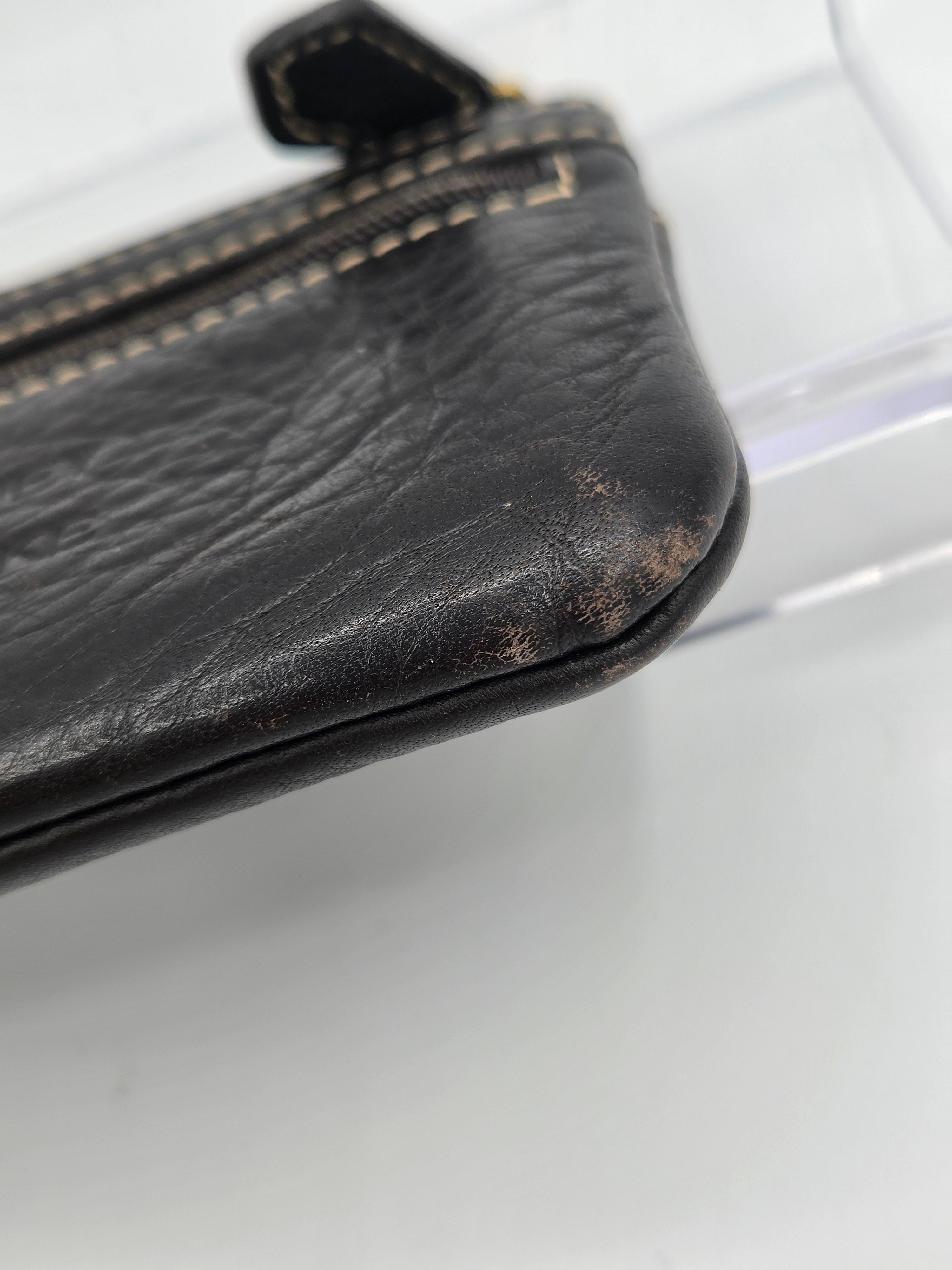 A close-up of a black distressed leather zippered pouch showing significant scuffing and wear on the bottom corner, with light beige contrast stitching.