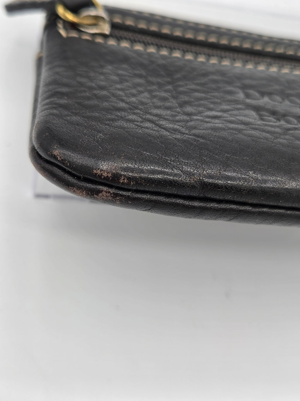 Close-up of a black pebbled leather zip pouch showing significant wear, scuffing, and tear marks along the bottom edge, with a gold-tone zipper pull.
