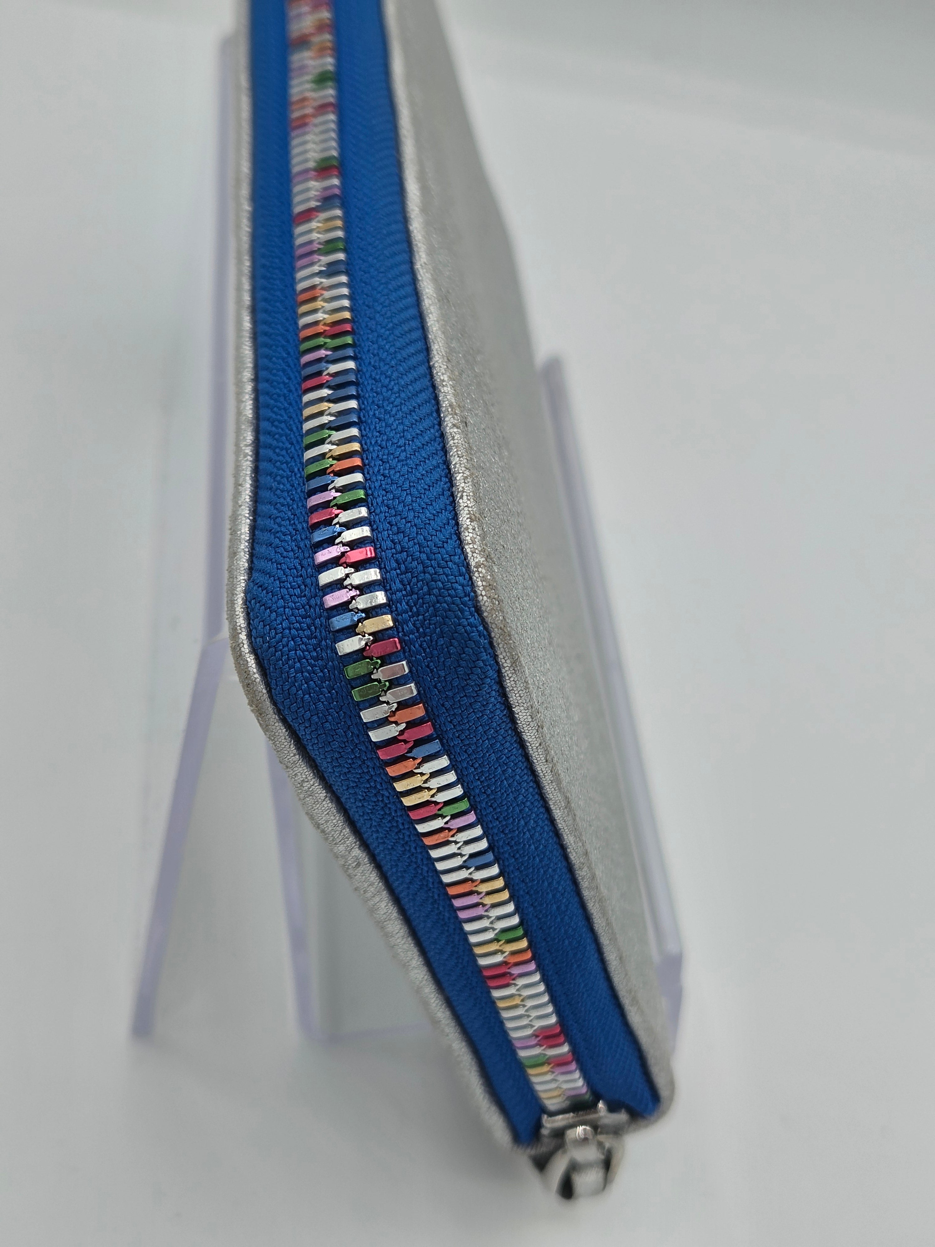 A close-up of a zippered pouch or wallet with a silver metallic exterior, a vibrant blue zipper track, and a decorative multicolor patterned accent running along the zipper.