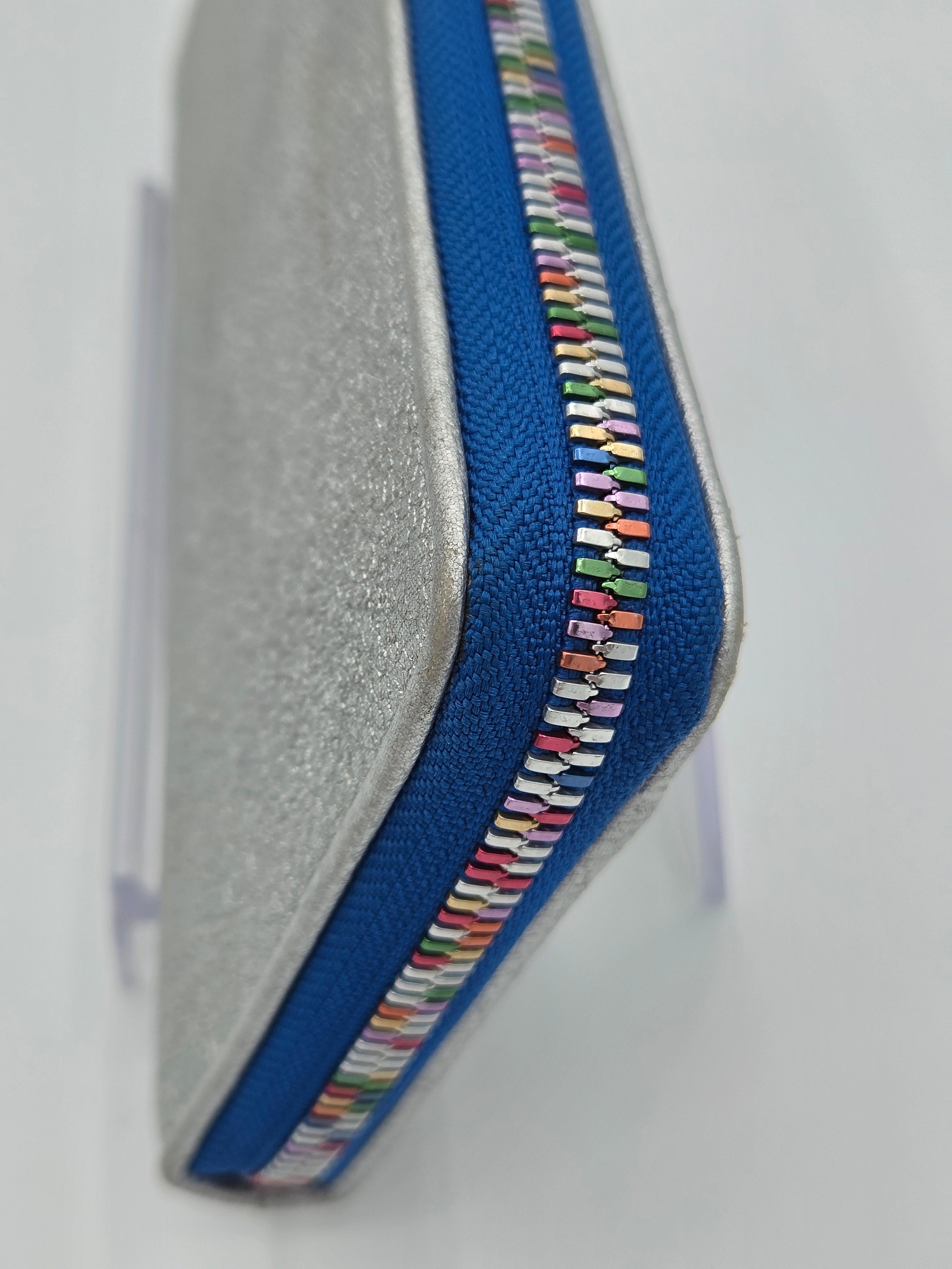 A close-up of a zippered pouch or case with a textured silver-gray exterior, a bright blue zipper track, and a decorative multicolor striped zipper pull detail, shown on a white background.