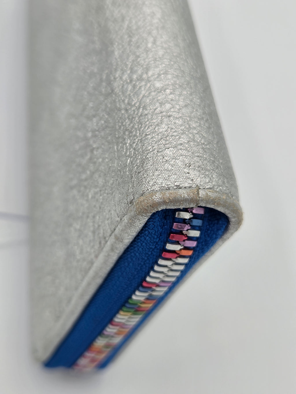 A close-up of a silver textured leather zippered sleeve or notebook cover, showing a blue fabric lining and a colorful array of small ribbon or thread loops along the zipper track, set against a plain white background.