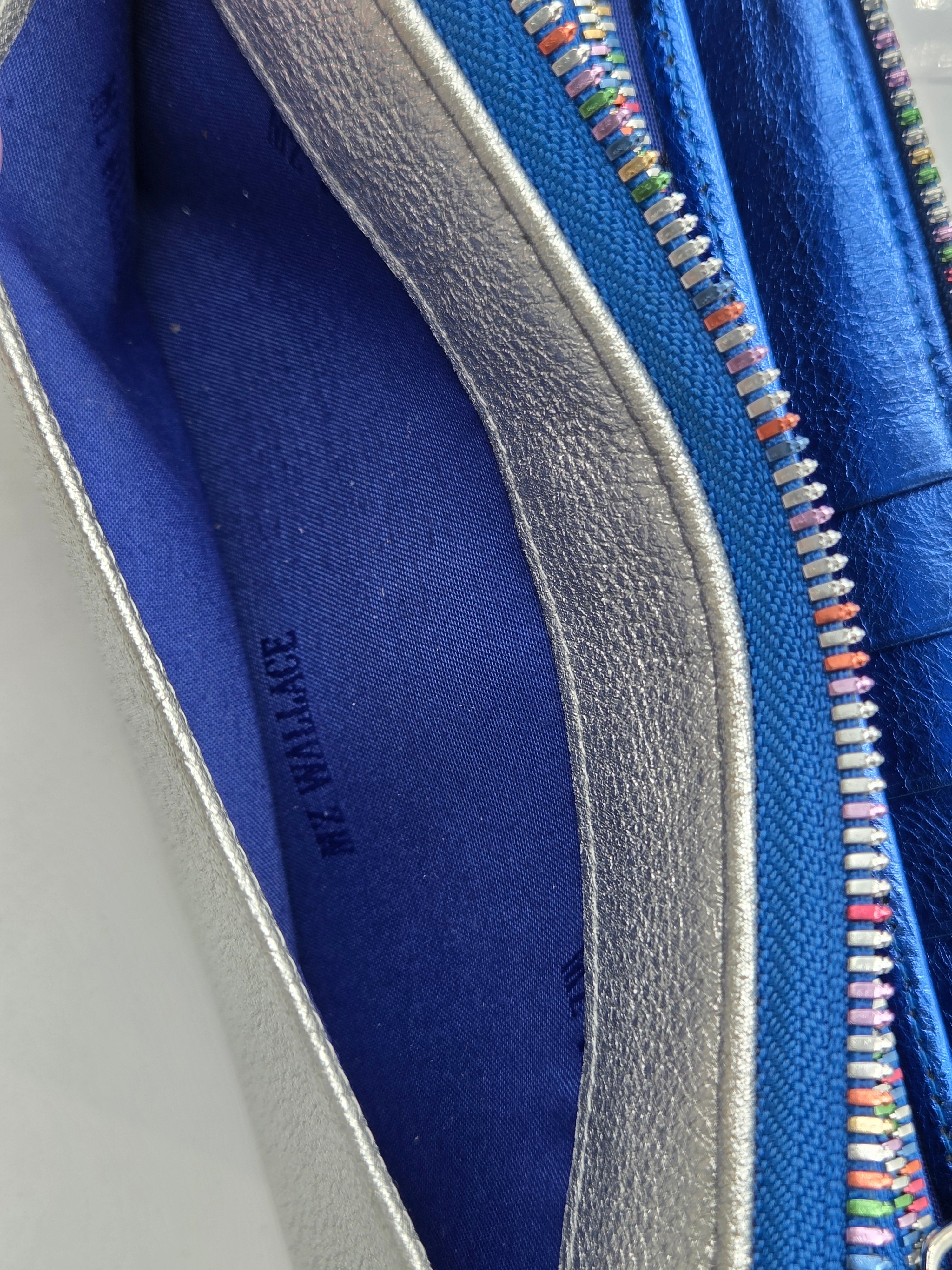 Close-up of a blue leather wallet or clutch showing the interior lining, silver-tone zipper, and multicolored zipper teeth, with 'M. MARCHAGE' printed inside.