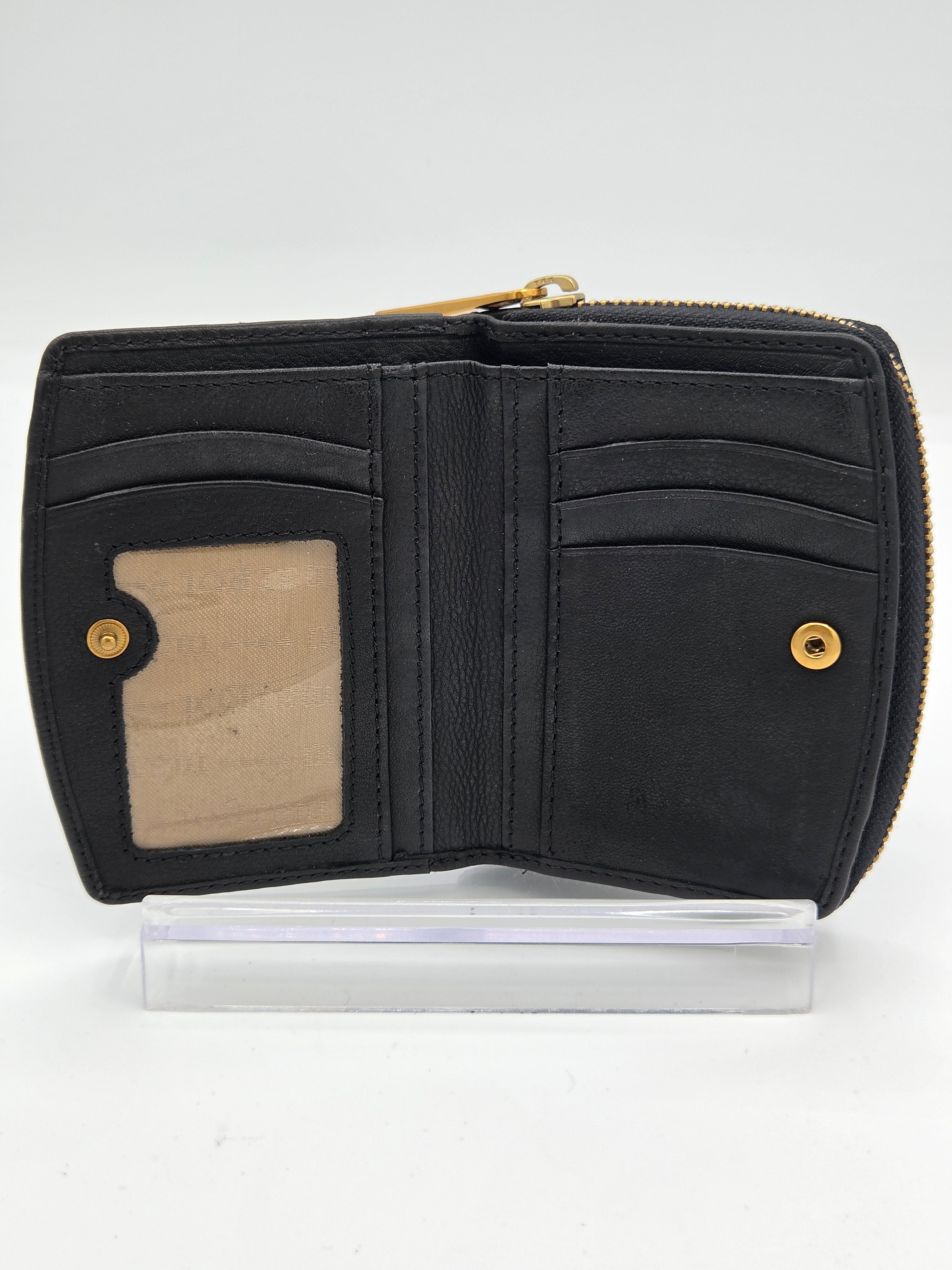 A black leather zip-around wallet shown open, displaying card slots, a clear ID window, and gold-tone hardware, resting on a clear plastic display block against a white background.