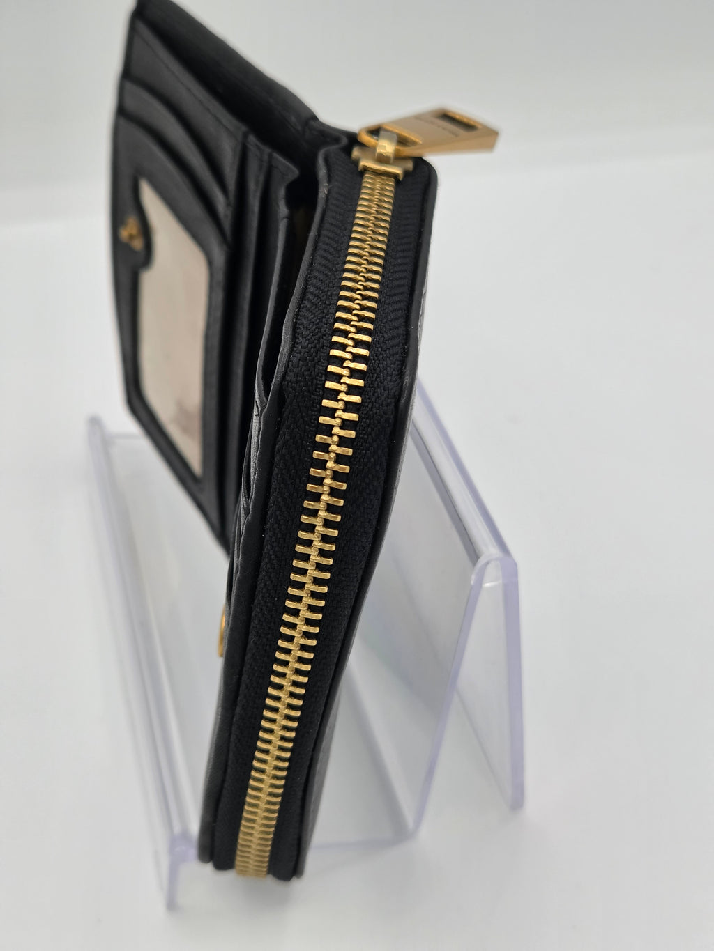 A black leather wallet with a gold zipper and gold hardware, displayed on a clear acrylic stand against a white background.