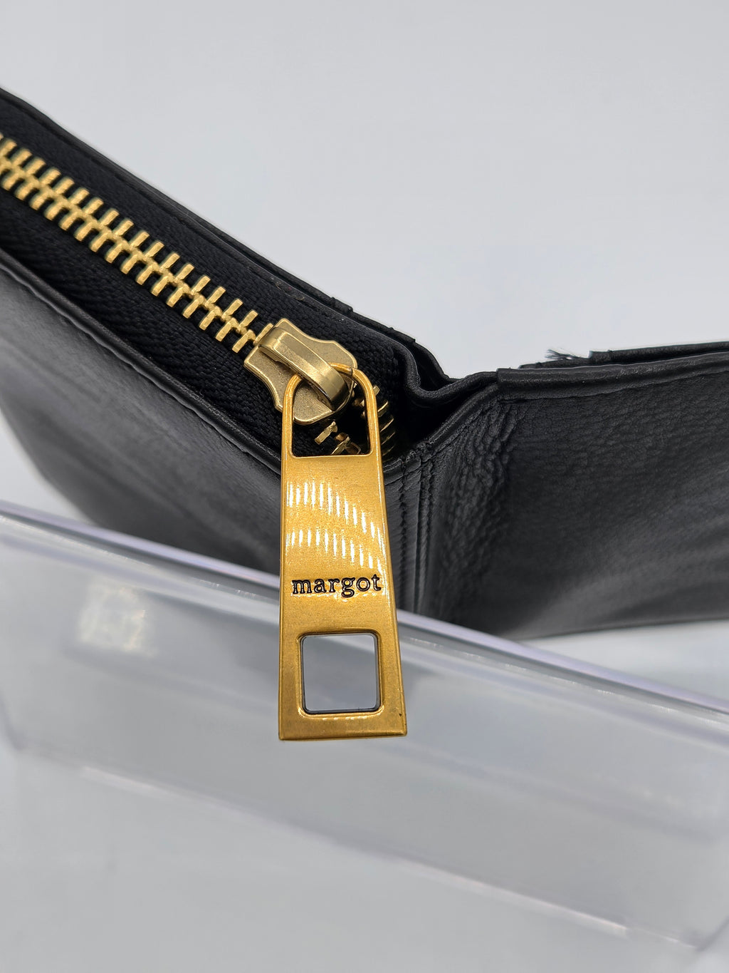 Close-up of a black leather zippered wallet or clutch with a prominent gold-tone metal zipper pull engraved with the brand name 'margot'.