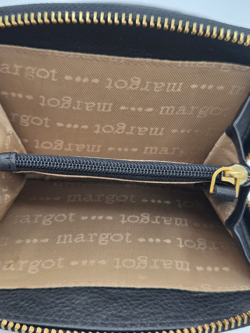 Close-up of the interior of a black leather bag with gold-tone zippers and a tan interior lining printed with the repeating 'margot' brand name.
