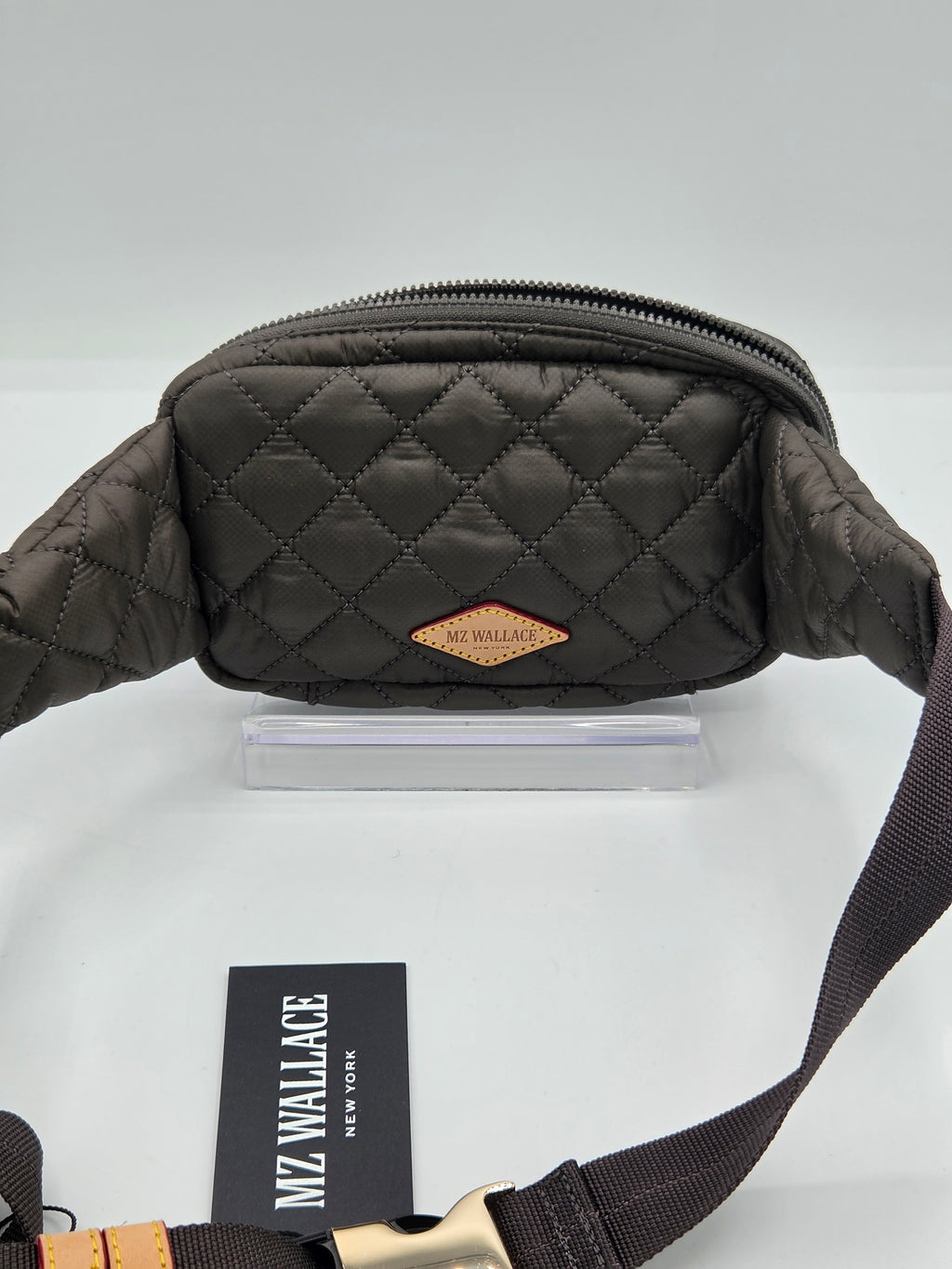A dark brown quilted fanny pack with a diamond-stitched pattern, a small tan leather brand patch, and a black adjustable strap, displayed on a white surface with a brand card.